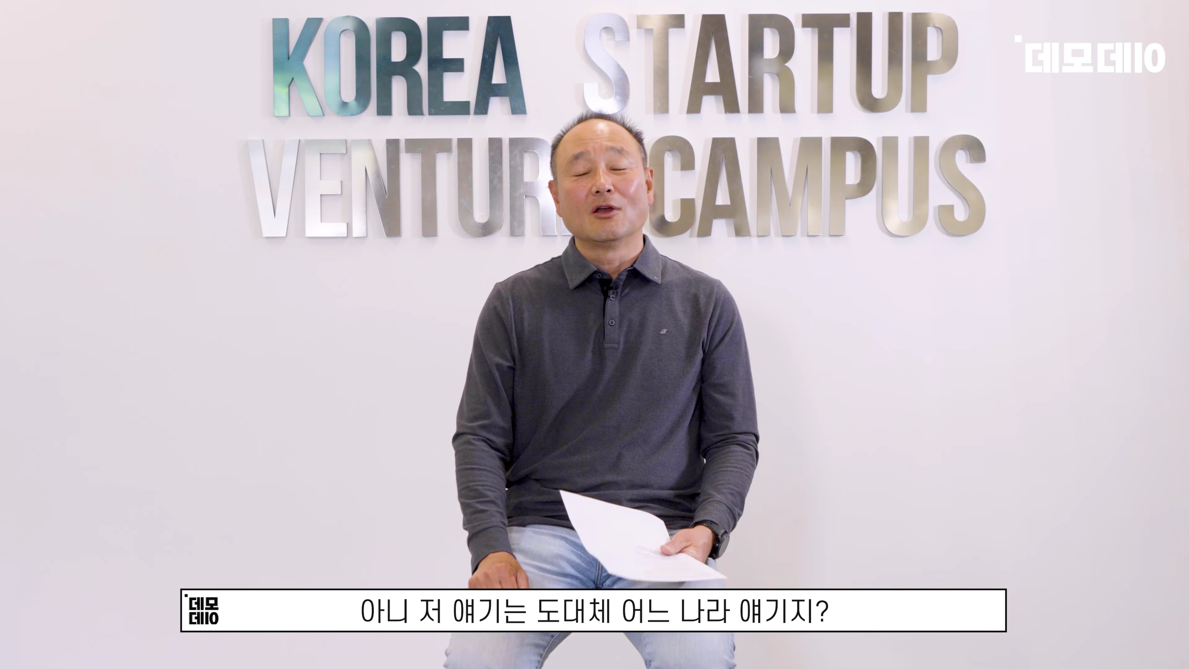 실내에서 회색 셔츠를 입은 중년 남성이 종이를 들고 앉아 인터뷰하는 모습