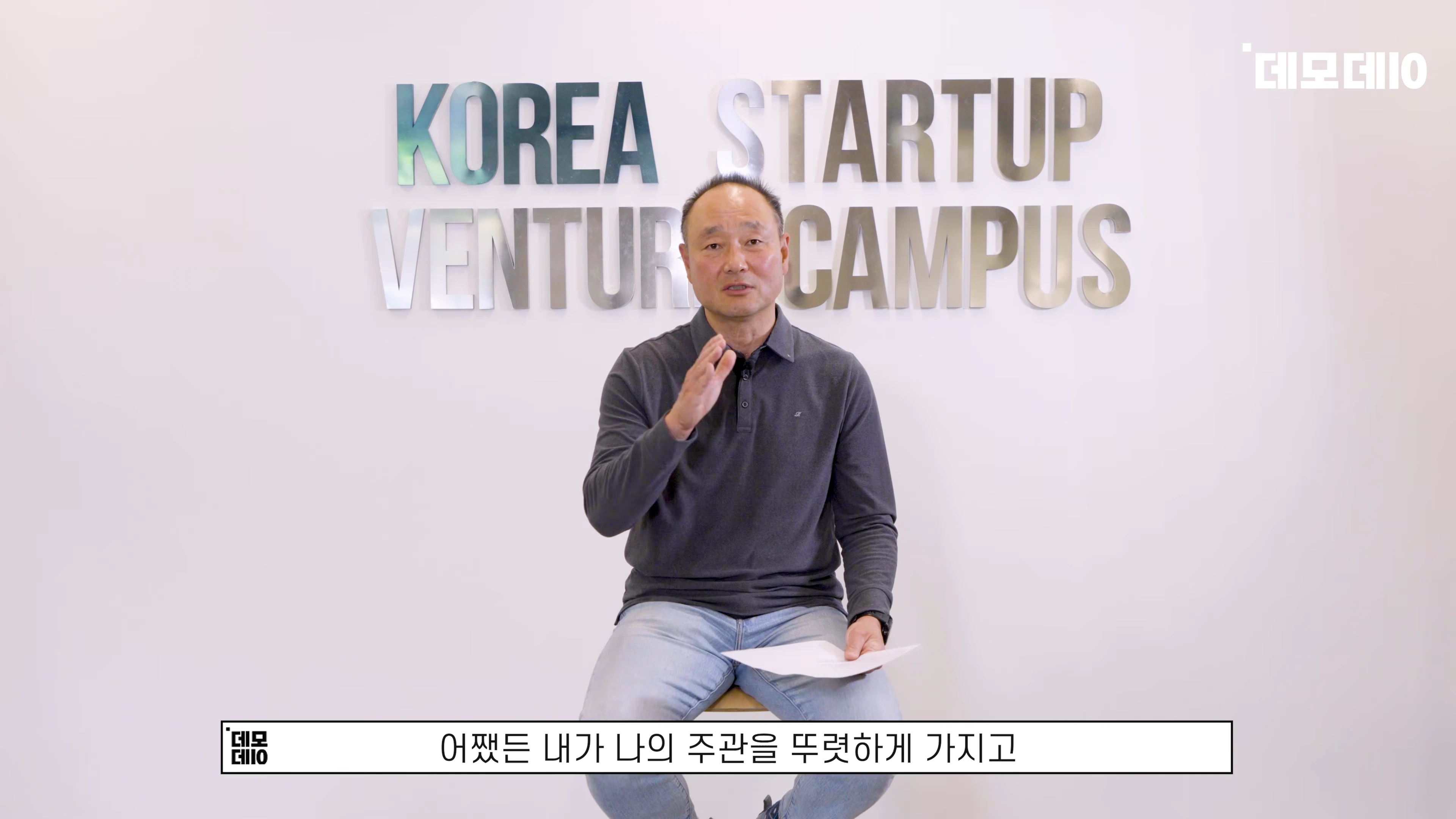 흰 벽을 배경으로 의자에 앉아 손짓하며 이야기하는 중년 남성