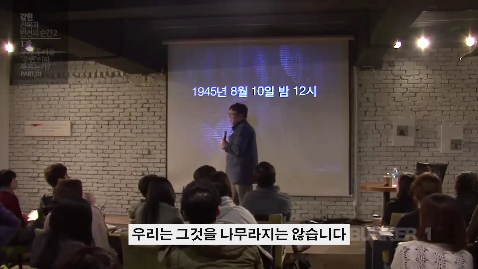 강연자가 스크린 앞에 서 있고 청중들이 앉아 있는 강연장 모습