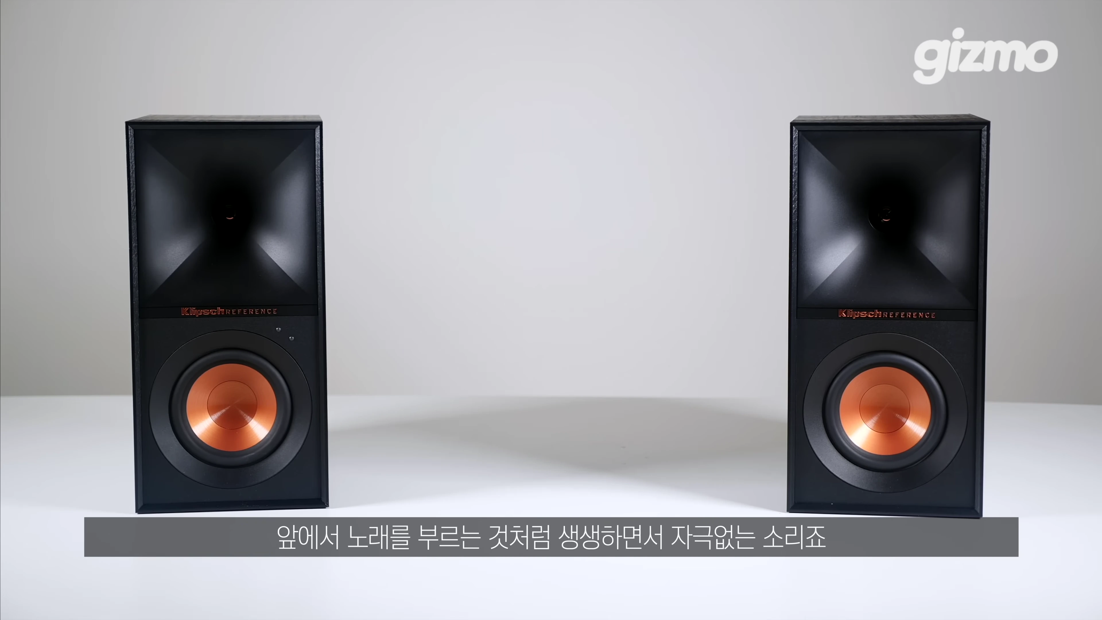 흰 배경 앞에 놓인 검은색 클립쉬 스피커 두 대와 하단에 자막이 표시된 영상 화면