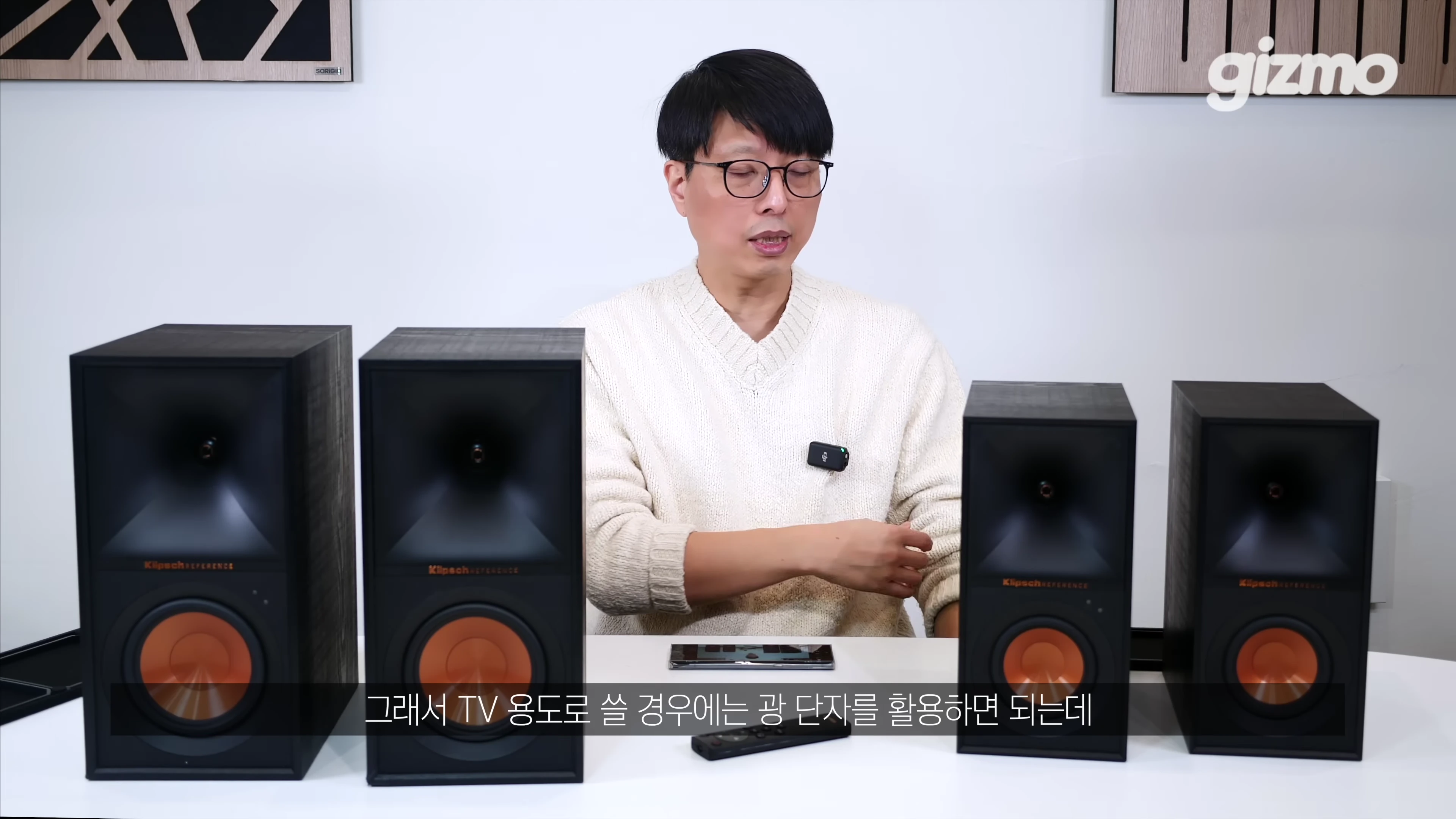 안경을 쓴 남성이 테이블 위에 놓인 두 쌍의 검은색 스피커 사이에서 설명하고 있다.