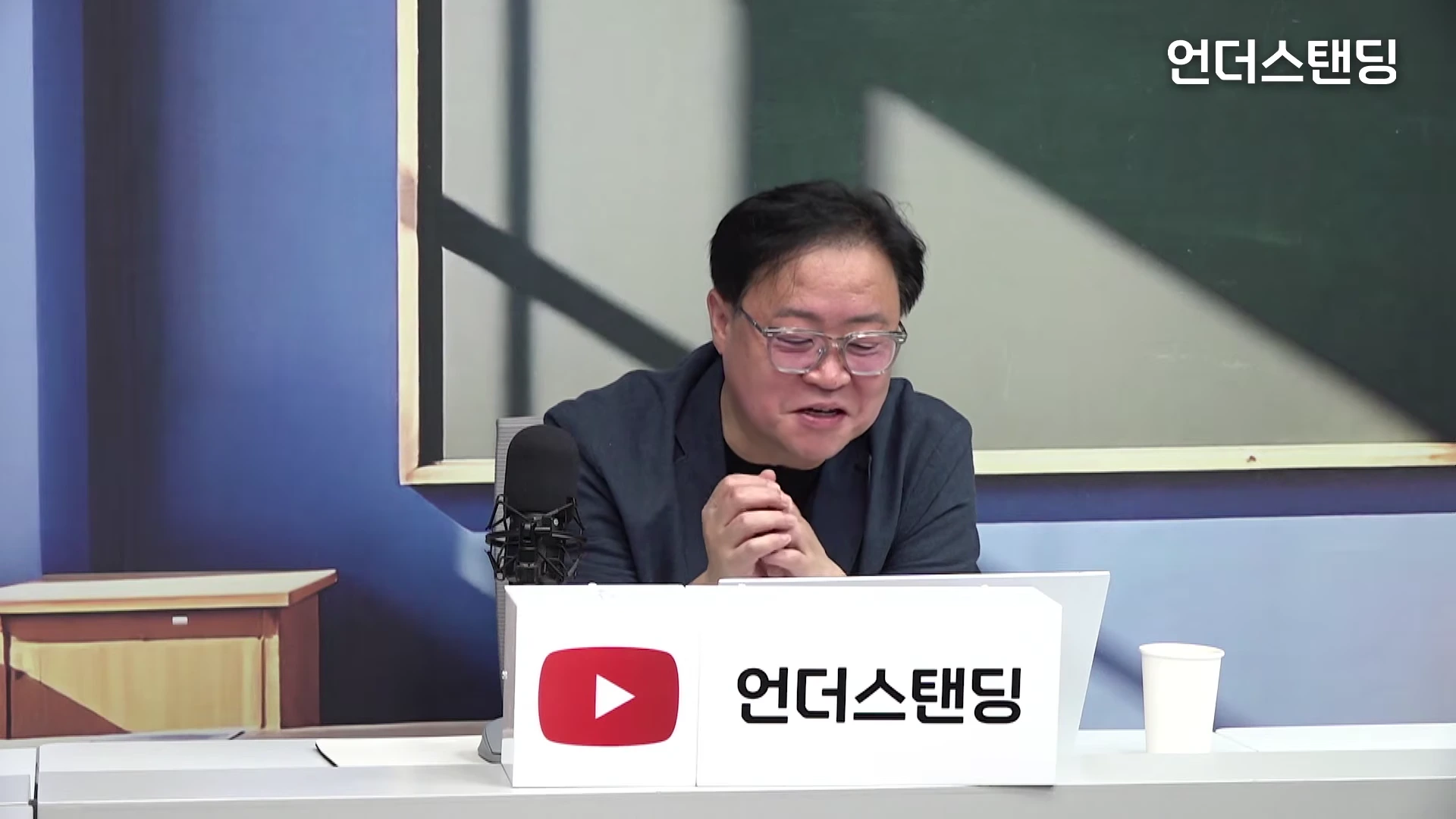 안경을 쓴 남성이 스튜디오 책상 앞에 앉아 이야기를 나누고 있다.