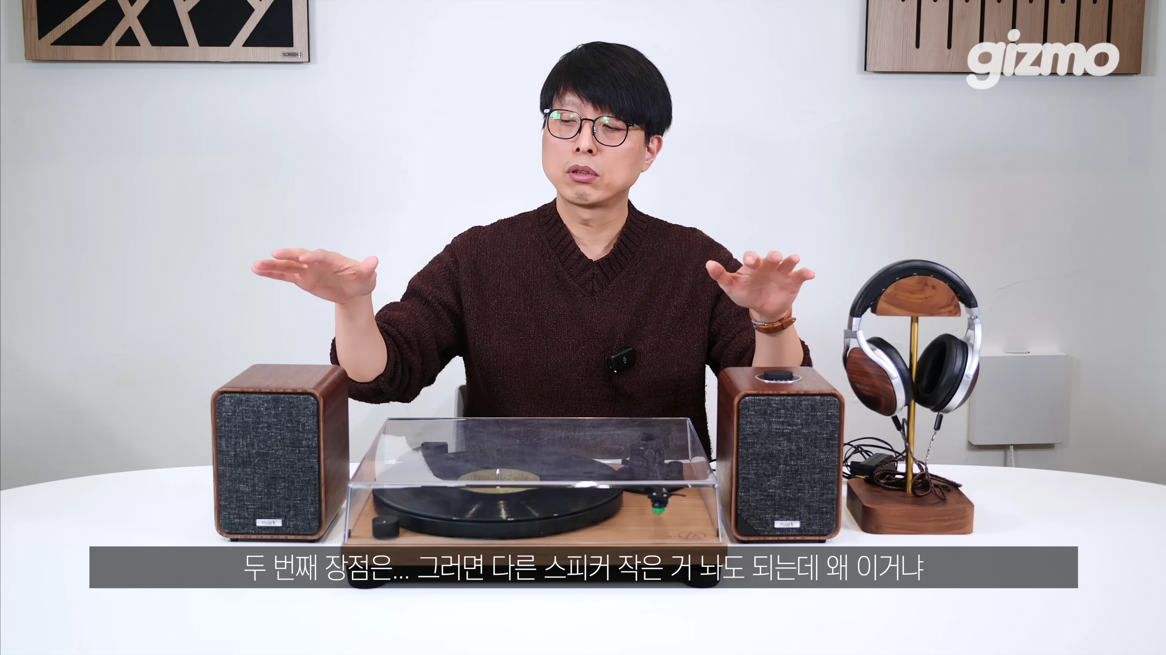 안경을 쓴 남성이 책상 위에 놓인 원목 스피커와 턴테이블을 가리키며 설명하고 있다.