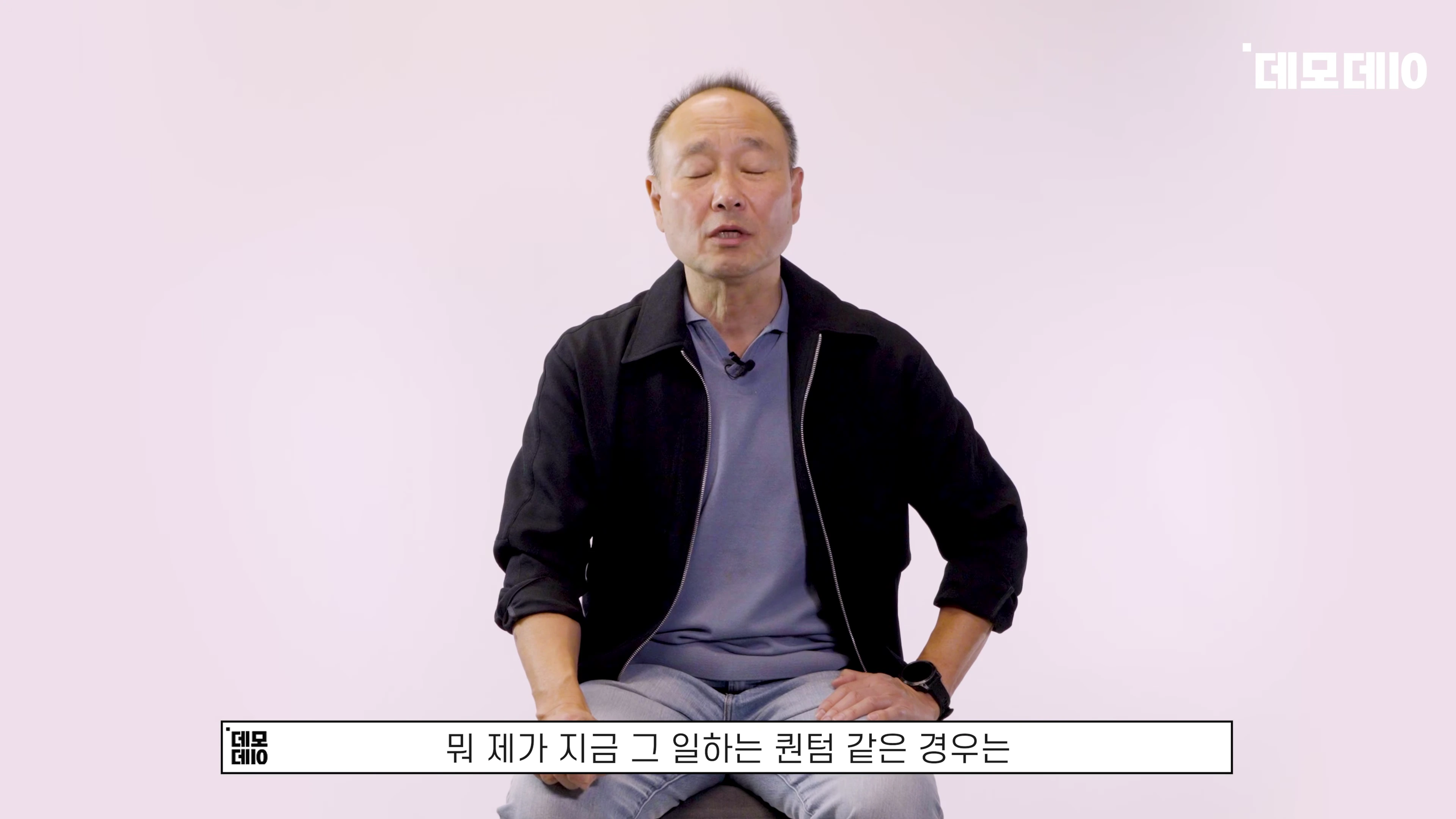밝은 배경 앞에서 검은 재킷을 입은 중년 남성이 앉아 인터뷰를 진행하고 있다.