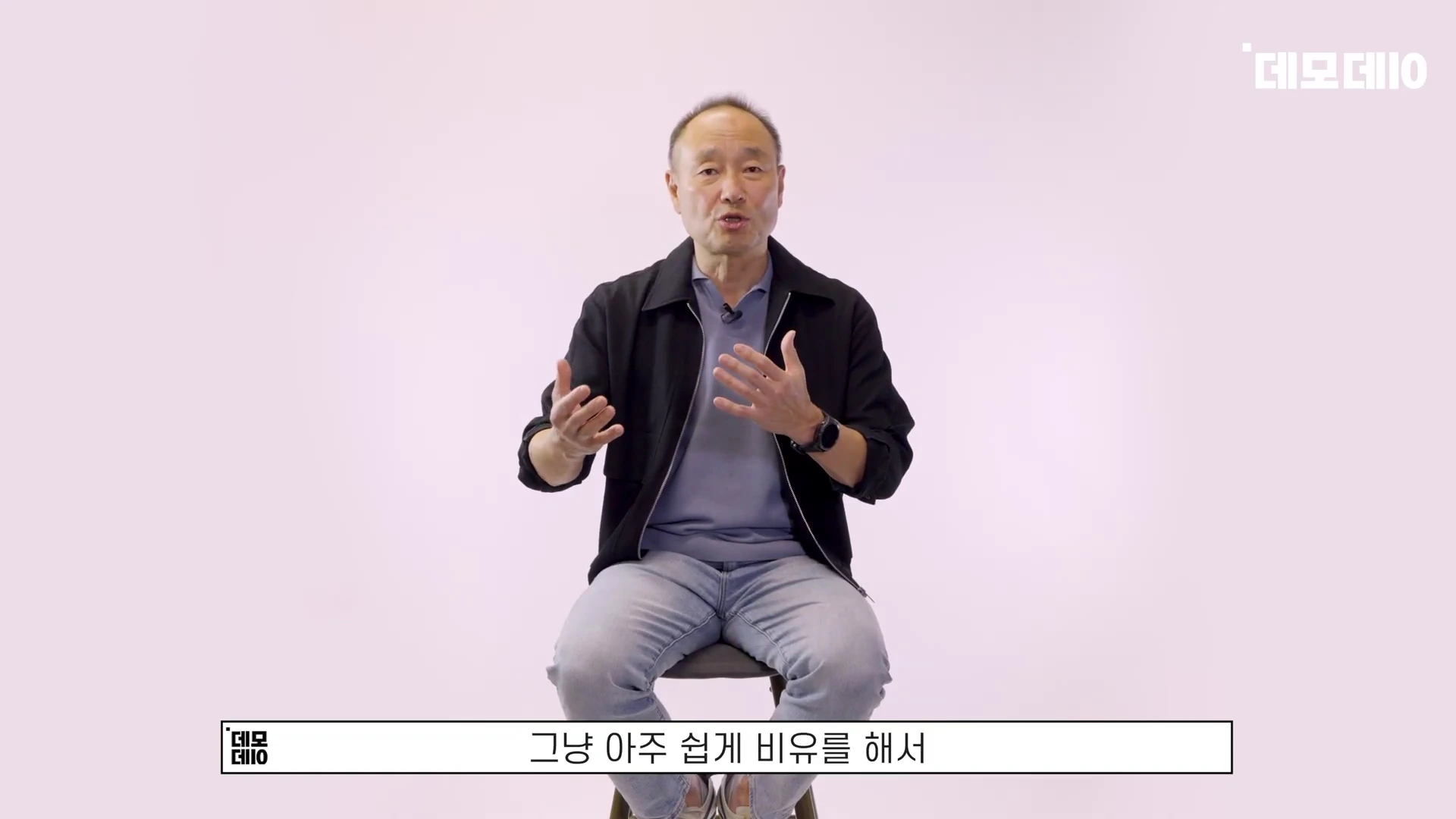흰 배경의 스튜디오에서 의자에 앉아 손짓하며 이야기하는 중년 남성
