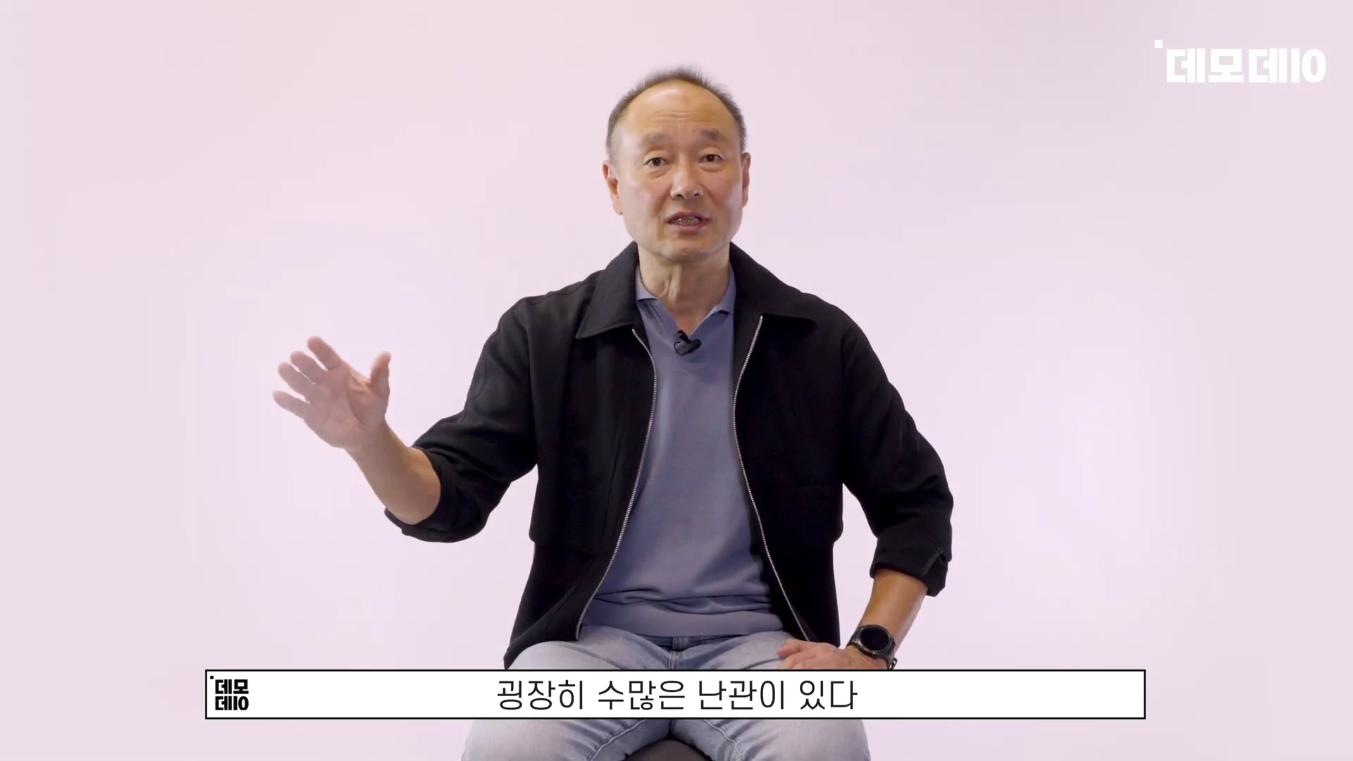 밝은 배경 앞에서 검은 재킷을 입은 중년 남성이 손을 들어 설명하고 있으며, 하단에는 자막이 표시되어 있다.