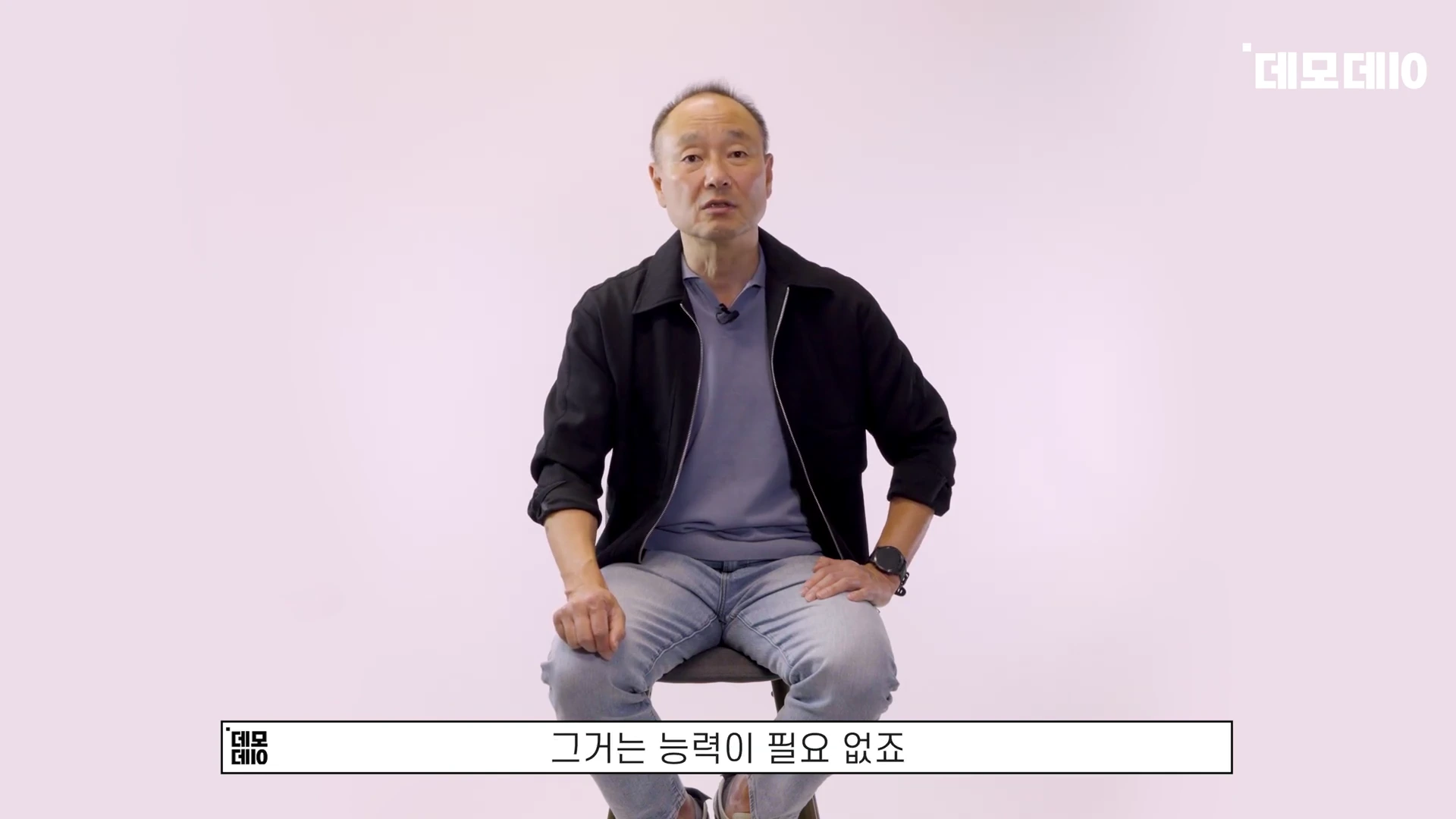 밝은 배경의 스튜디오 의자에 앉아 정면을 응시하며 이야기하는 중년 남성