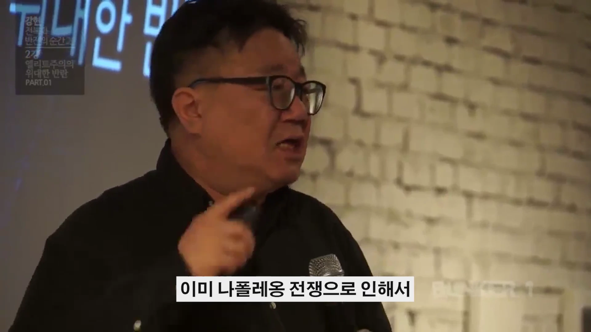 안경을 쓴 중년 남성이 강연장에서 마이크를 들고 열정적으로 이야기하고 있다.
