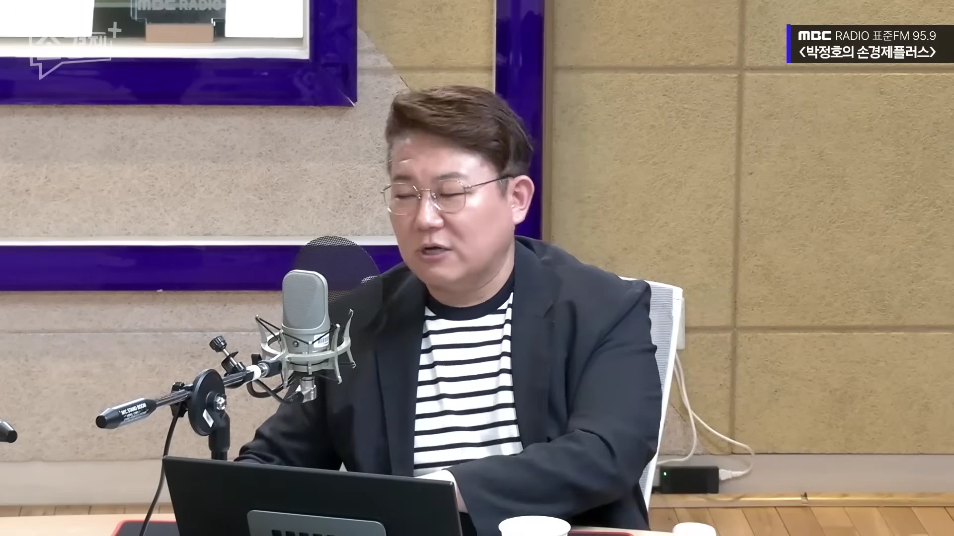 안경을 쓴 남성이 라디오 스튜디오에서 마이크 앞에 앉아 이야기를 나누고 있다.