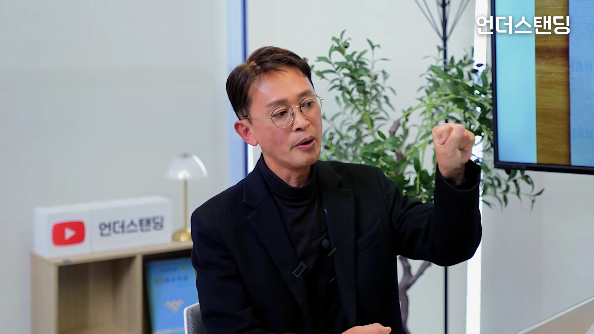 안경을 쓴 남성이 스튜디오에서 손을 들어 보이며 열정적으로 이야기하고 있다.