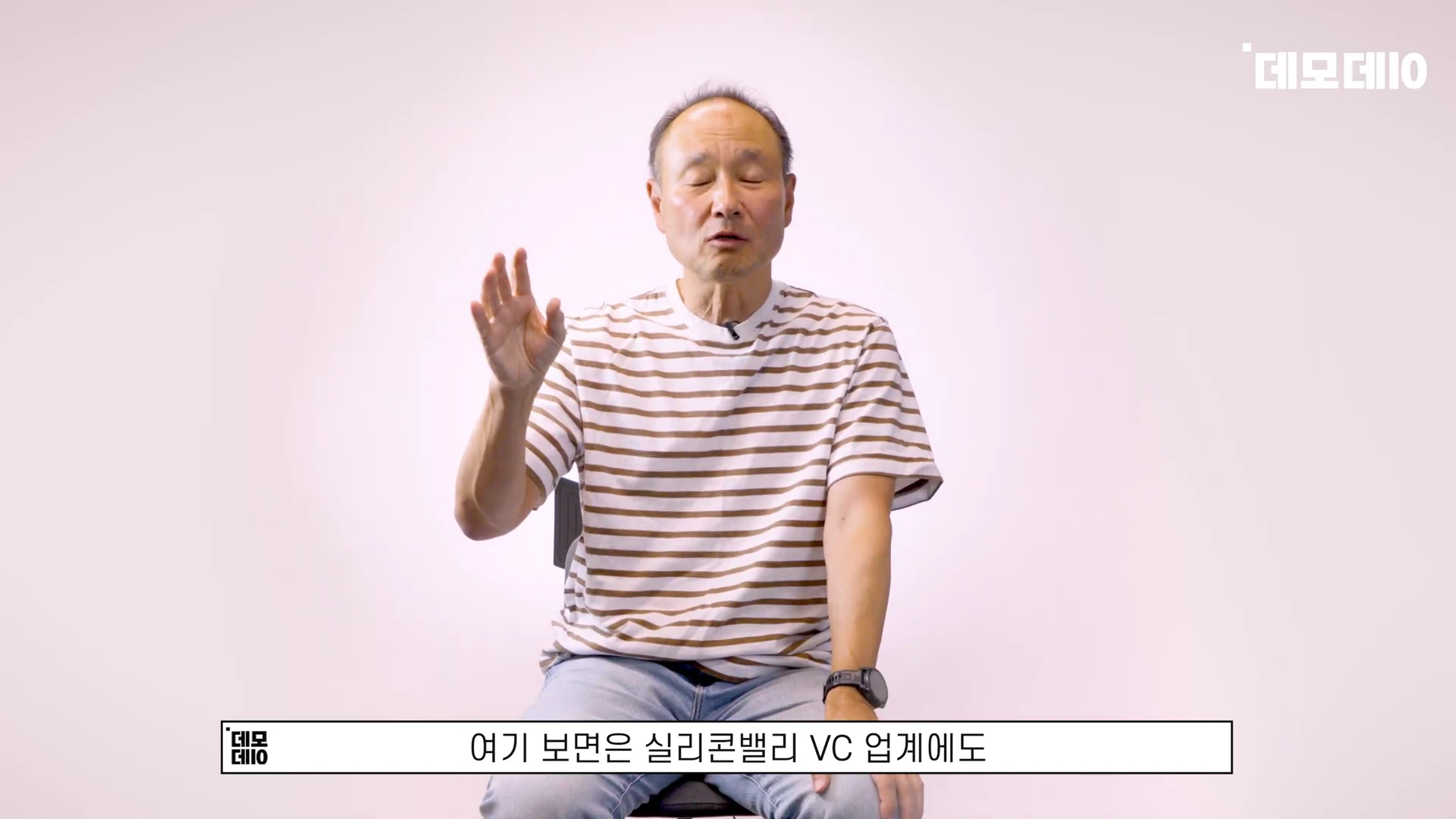 줄무늬 티셔츠를 입은 중년 남성이 밝은 배경 앞에서 손을 들어 대화하고 있는 모습