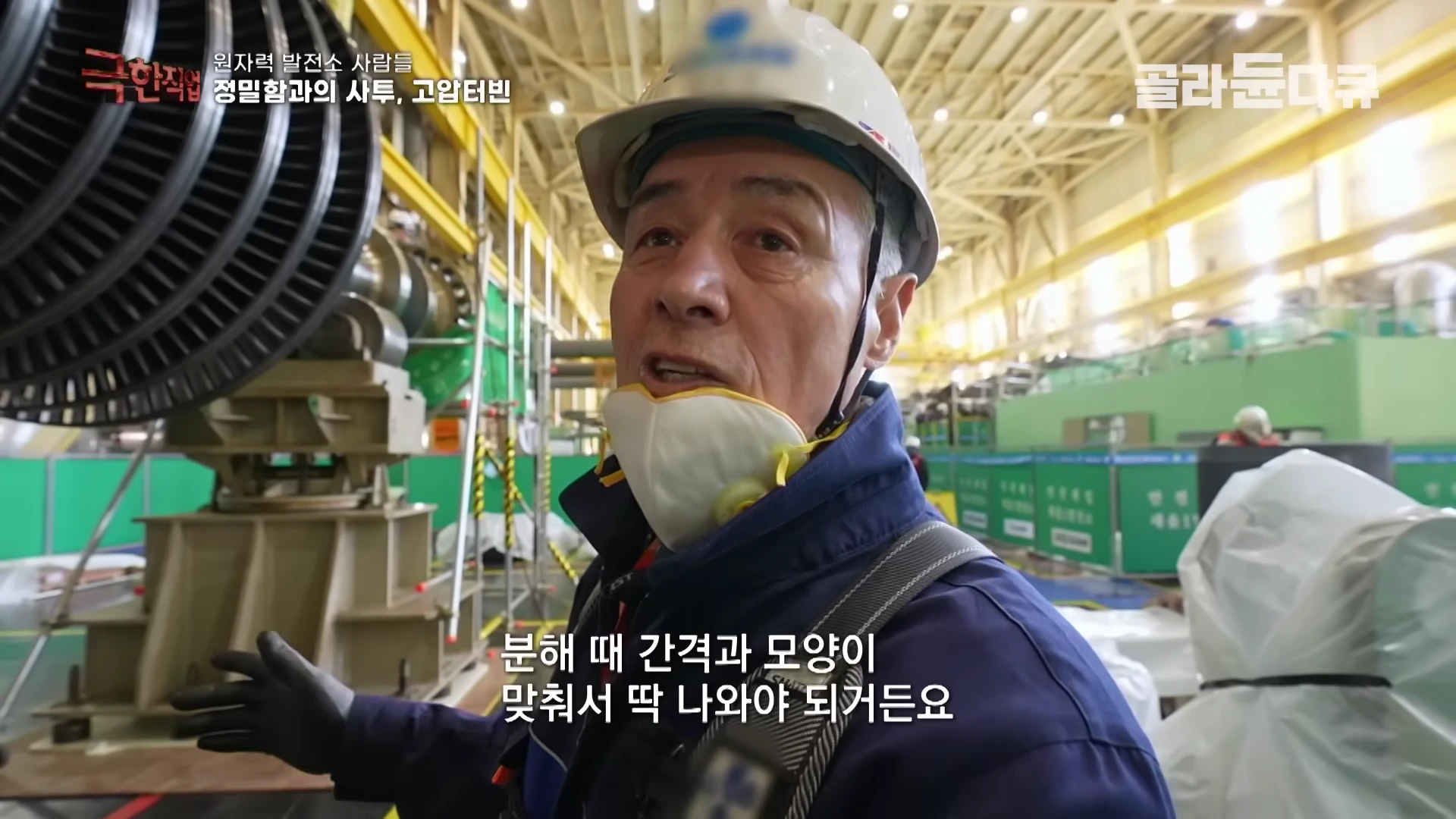 안전모와 마스크를 착용한 작업자가 원자력 발전소 내부에서 터빈 부품을 배경으로 인터뷰하고 있다.