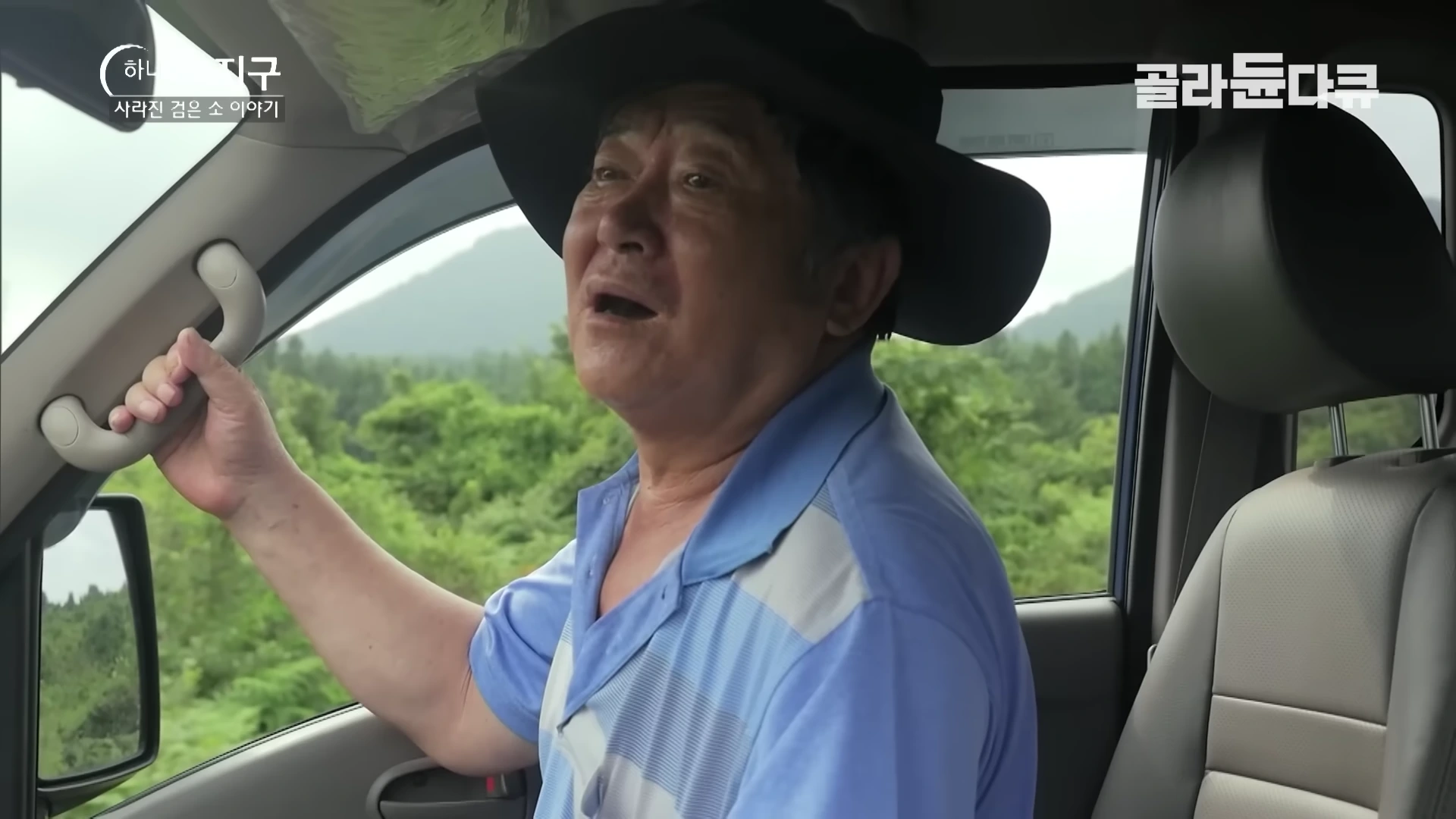 검은 모자를 쓴 중년 남성이 자동차 조수석에 앉아 밖을 보며 노래를 부르는 모습