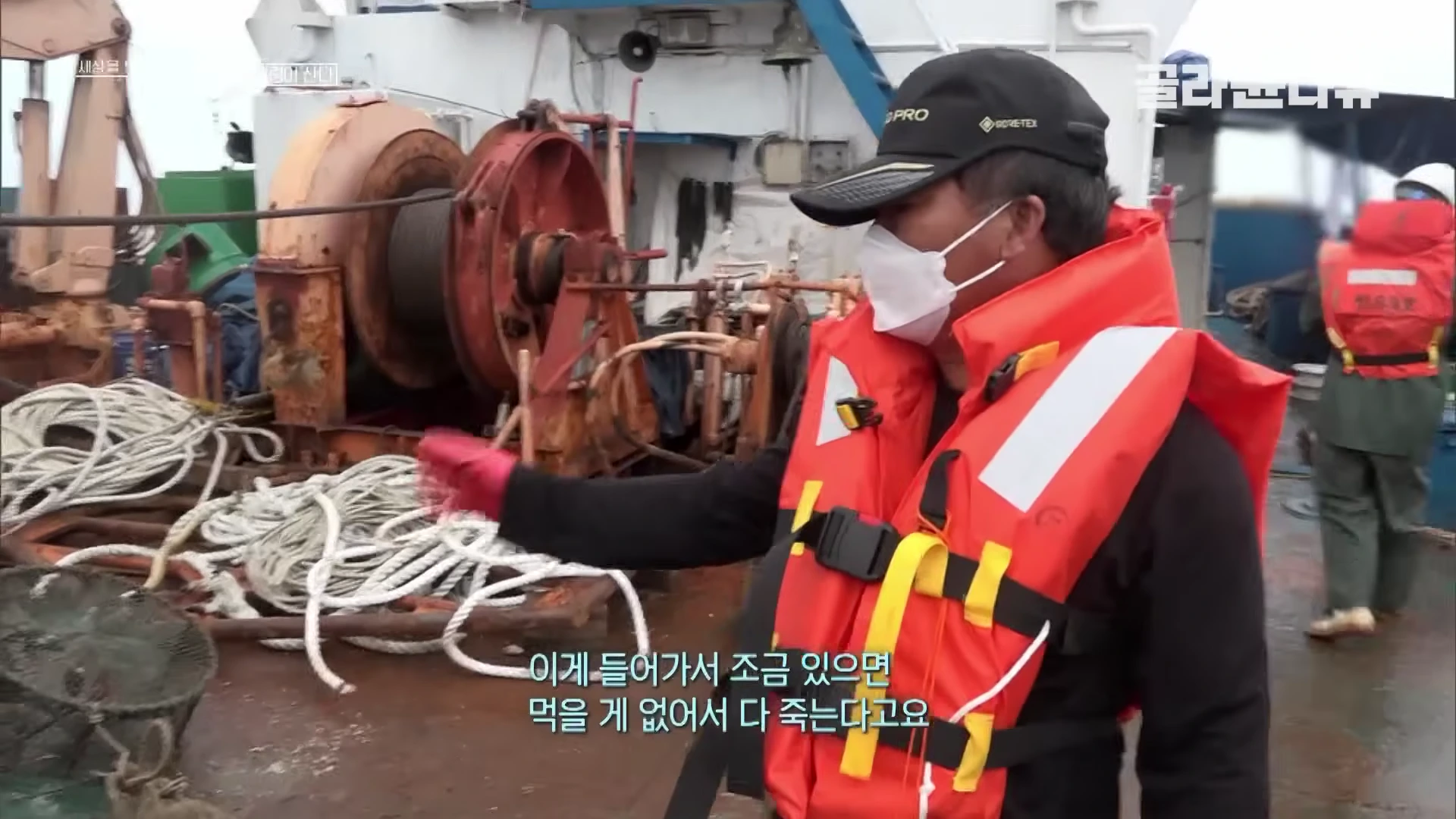 어선 갑판 위에서 주황색 구명조끼를 입은 남성이 폐어구 더미를 가리키며 설명하고 있다.