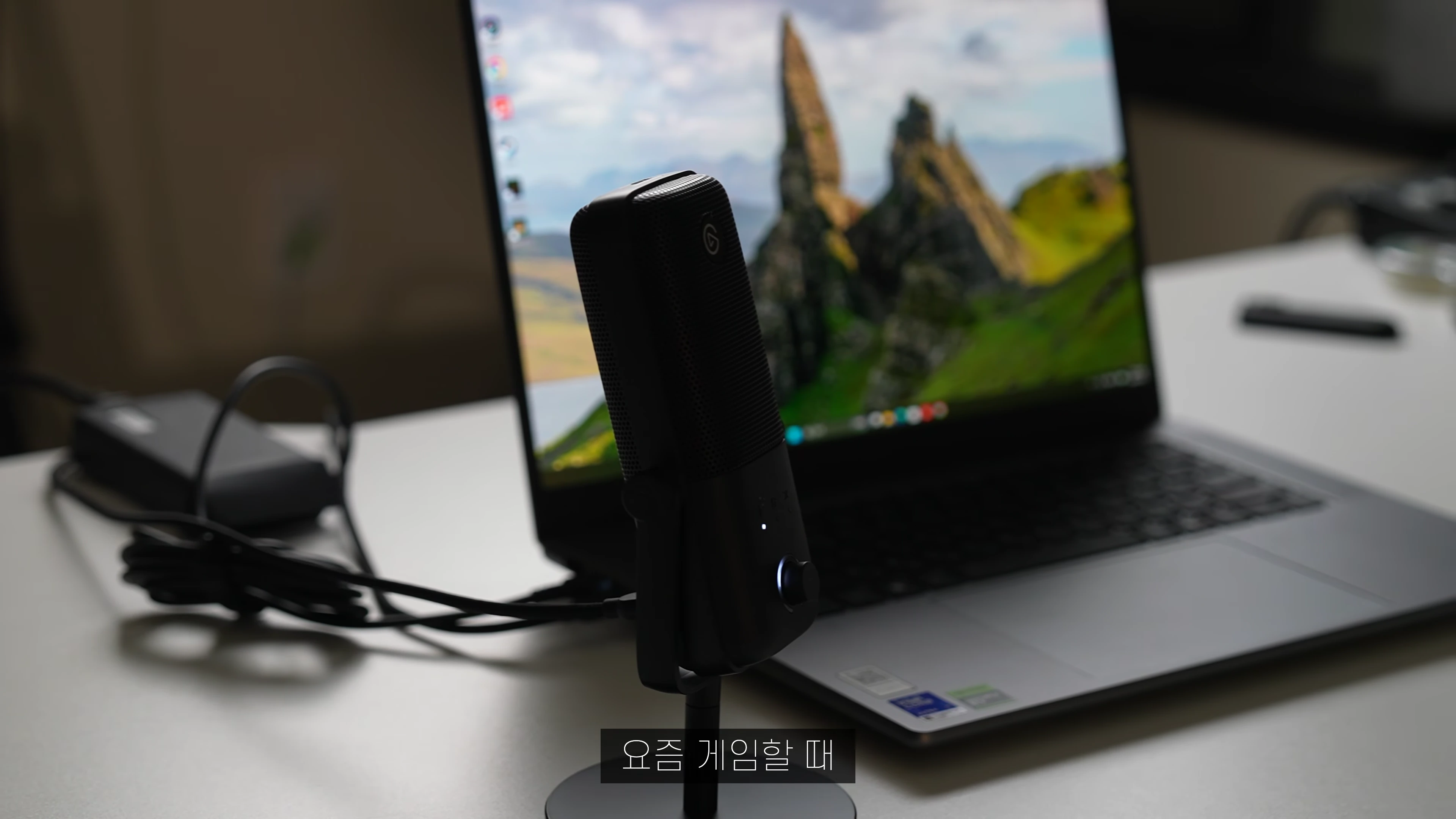 책상 위에 놓인 노트북 앞에 검은색 USB 마이크가 스탠드에 세워져 있는 모습