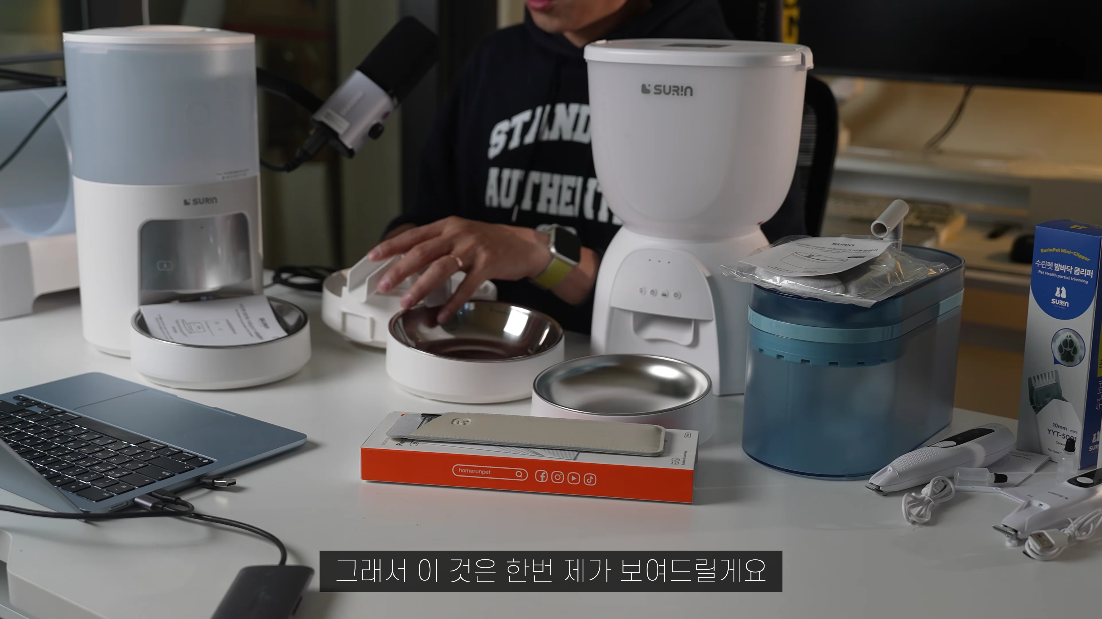 책상 위에 놓인 수린펫 자동급식기 본체와 듀얼 급식기용 액세서리, 그리고 발바닥 미용기 패키지
