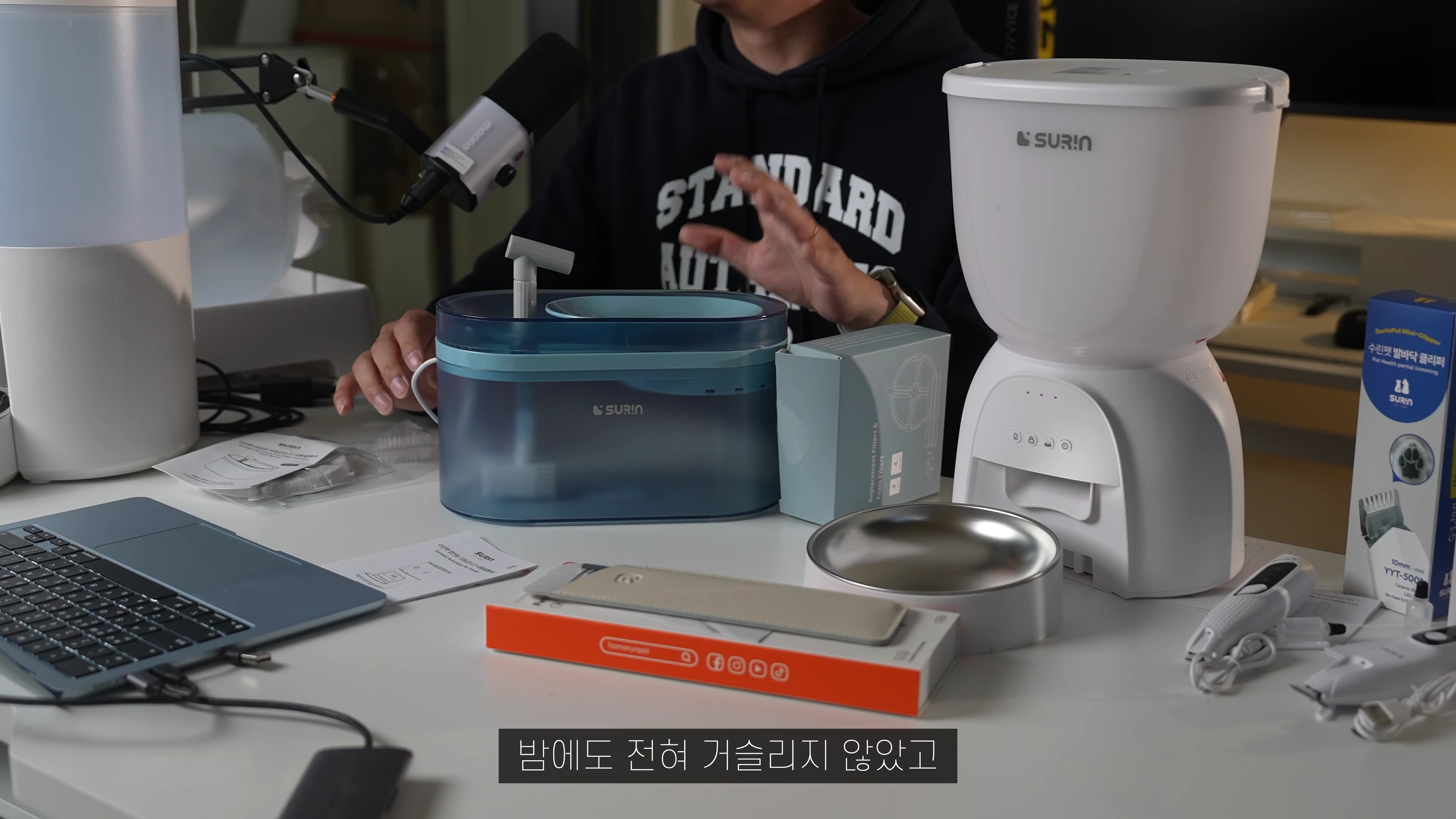 책상 위에 자동급식기, 자동급수기, 미용기 등 반려동물 용품이 놓여 있고 한 사람이 손짓하며 설명하는 모습