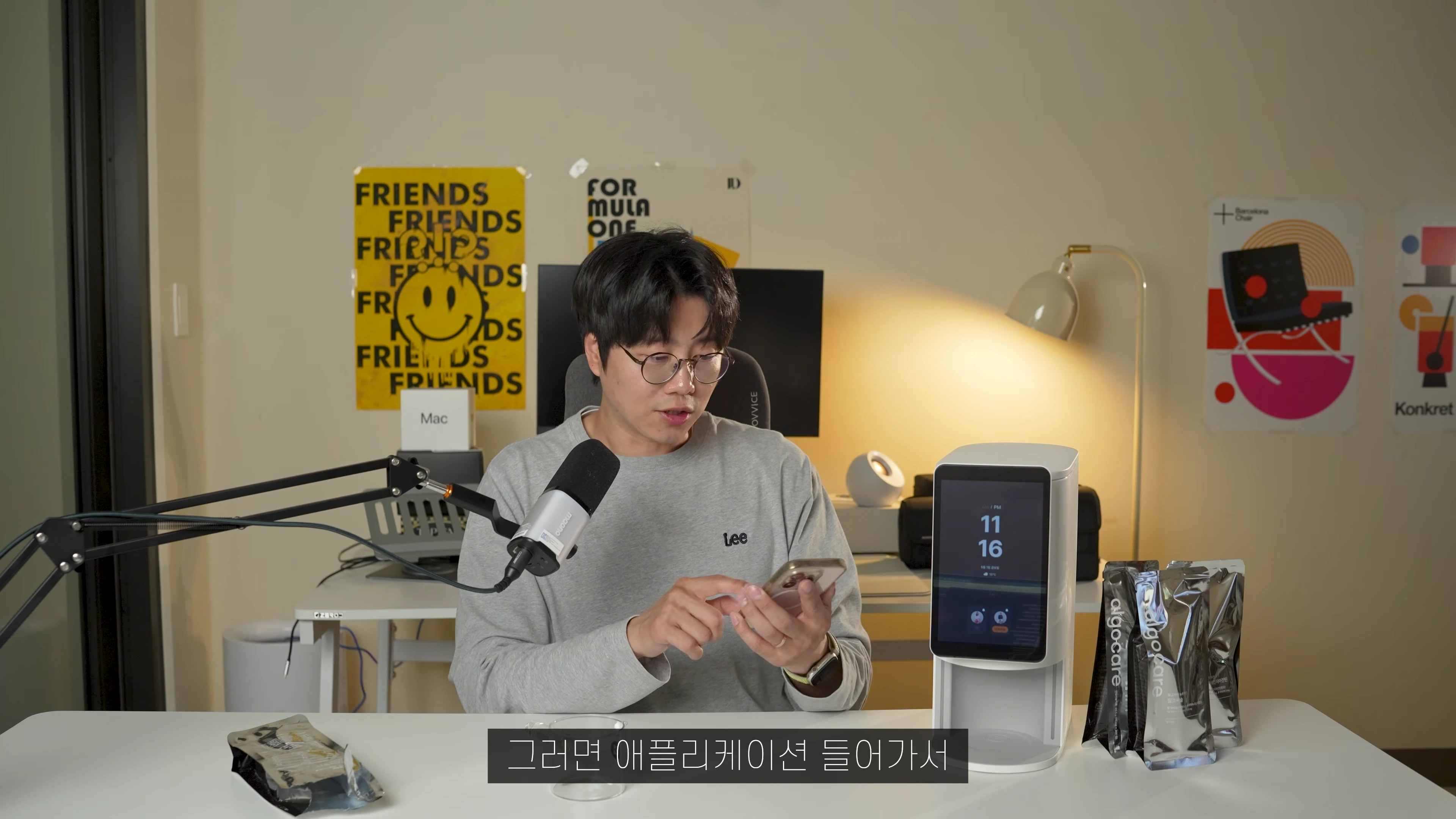 안경을 쓴 남성이 책상에 앉아 스마트폰을 보며 영양제 기기를 조작하고 있다.