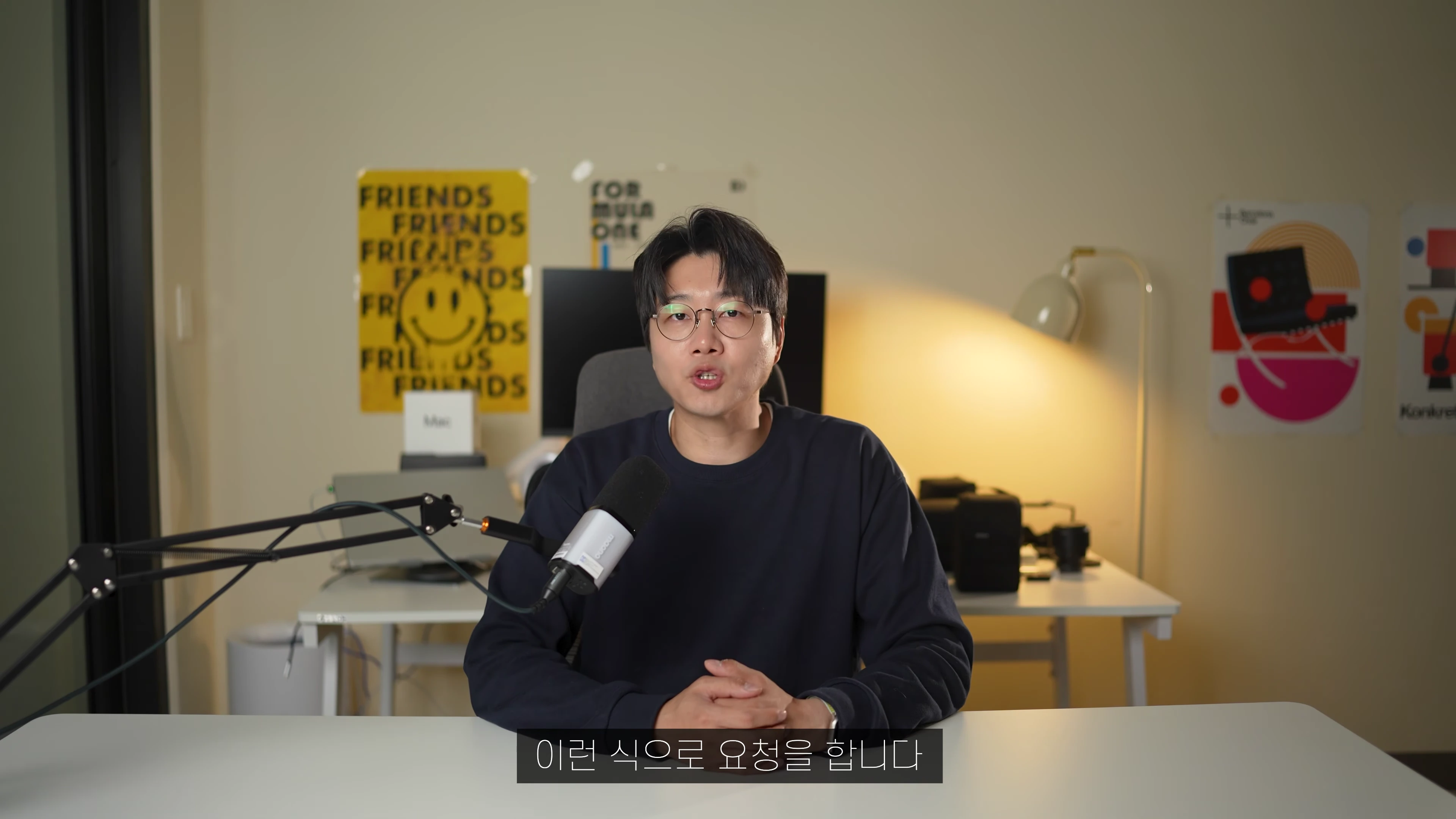 안경을 쓴 남성이 책상 앞에 앉아 마이크를 앞에 두고 이야기하는 모습