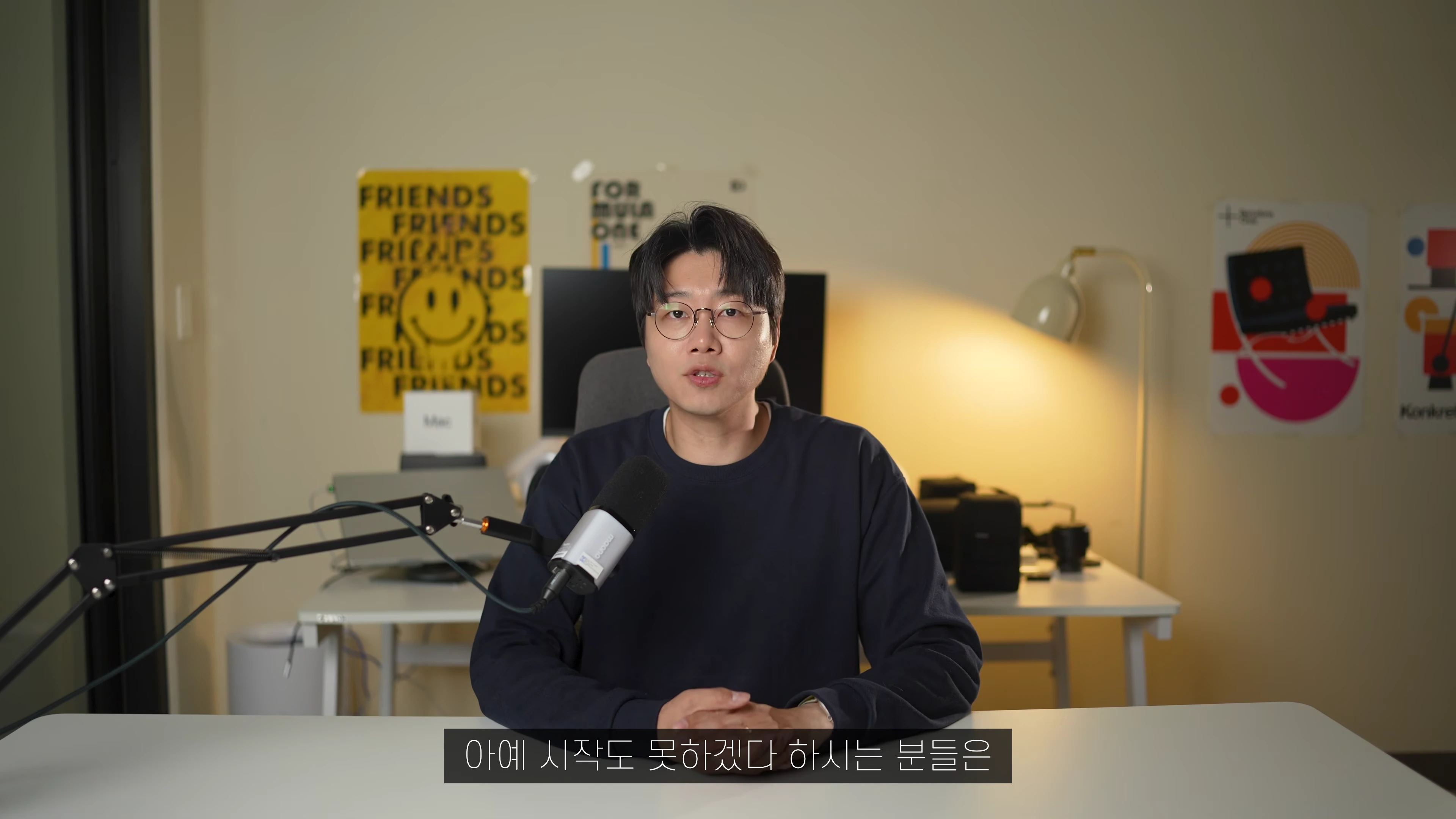 안경을 쓴 남성이 책상 앞에 앉아 마이크를 앞에 두고 이야기하는 모습