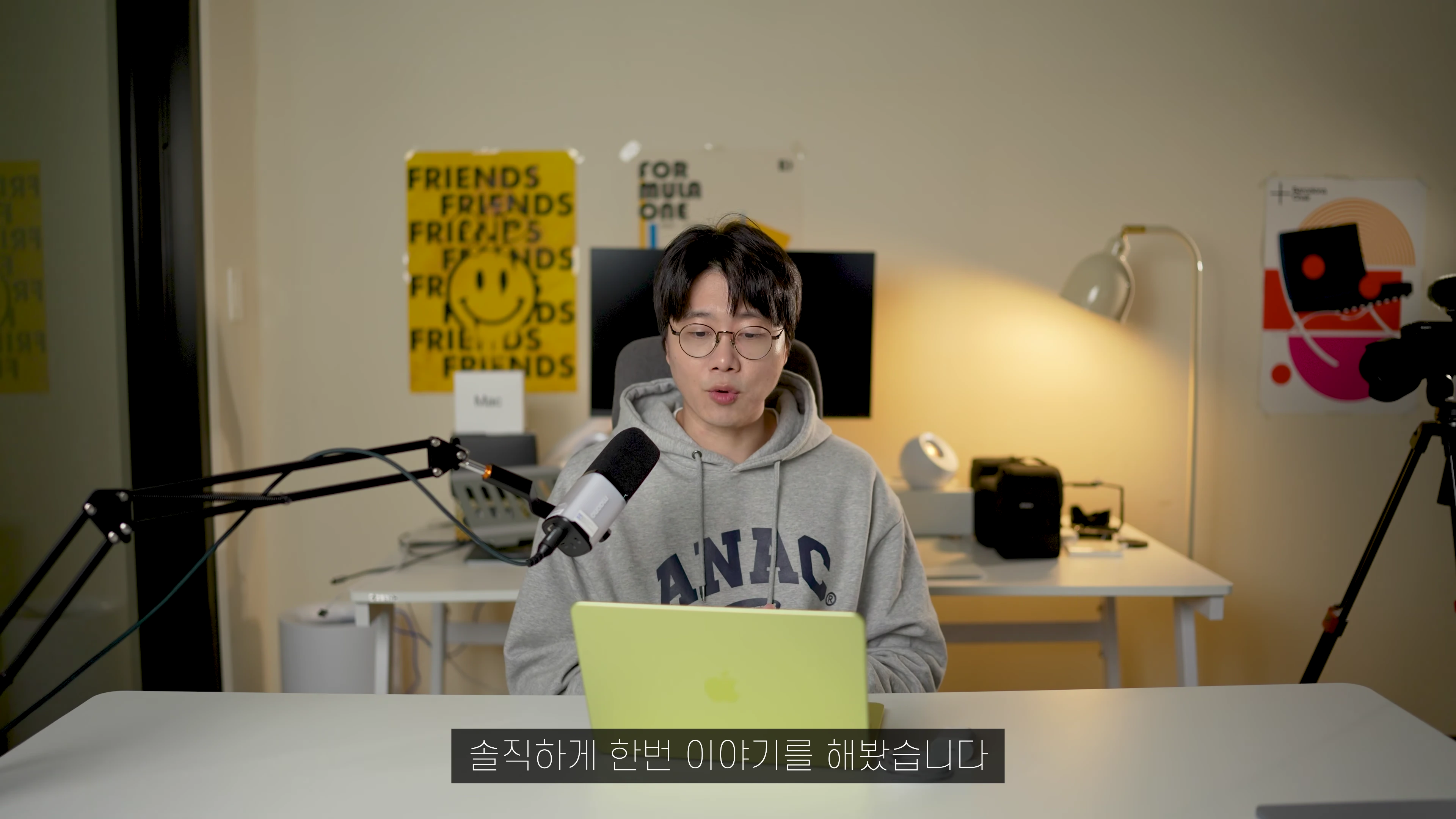 안경을 쓴 남성이 책상에 앉아 라임색 노트북을 사용하며 마이크를 앞에 두고 이야기하는 모습