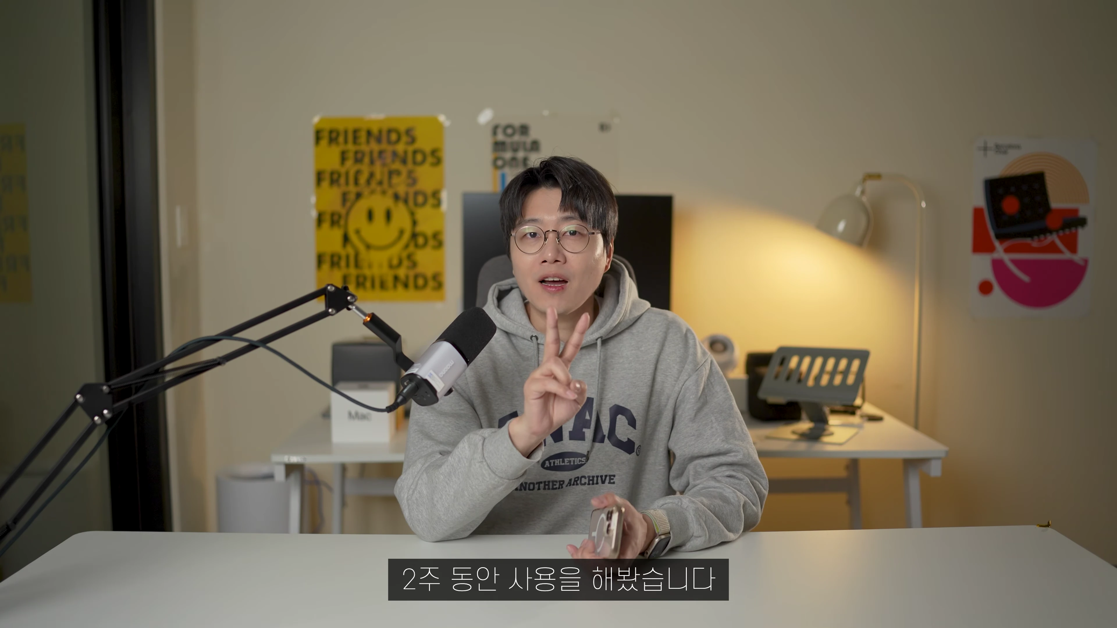 안경을 쓴 남성이 책상 앞에 앉아 손가락으로 숫자 2를 표시하며 이야기하고 있다.