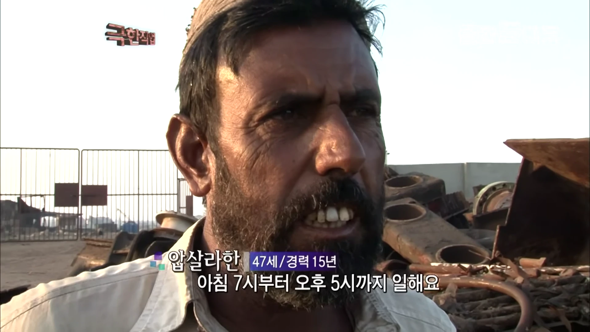 수염이 덥수룩한 중년 남성이 인터뷰하는 모습으로, 배경에는 철거 현장의 고철들이 쌓여 있다.