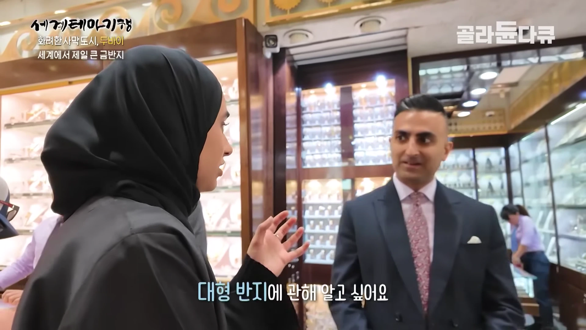 검은 히잡을 쓴 여성과 정장 차림의 남성이 금은방 안에서 대화를 나누고 있다.
