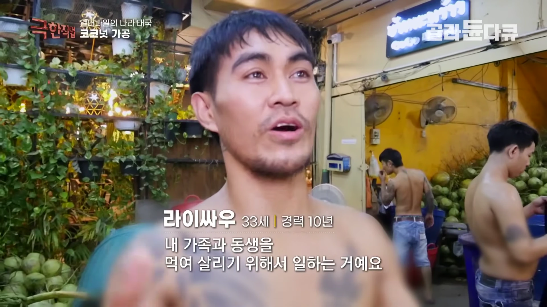 태국 코코넛 가공 작업장에서 상의를 탈의한 남성이 인터뷰하고 있으며, 뒤편으로 코코넛이 쌓여 있다.
