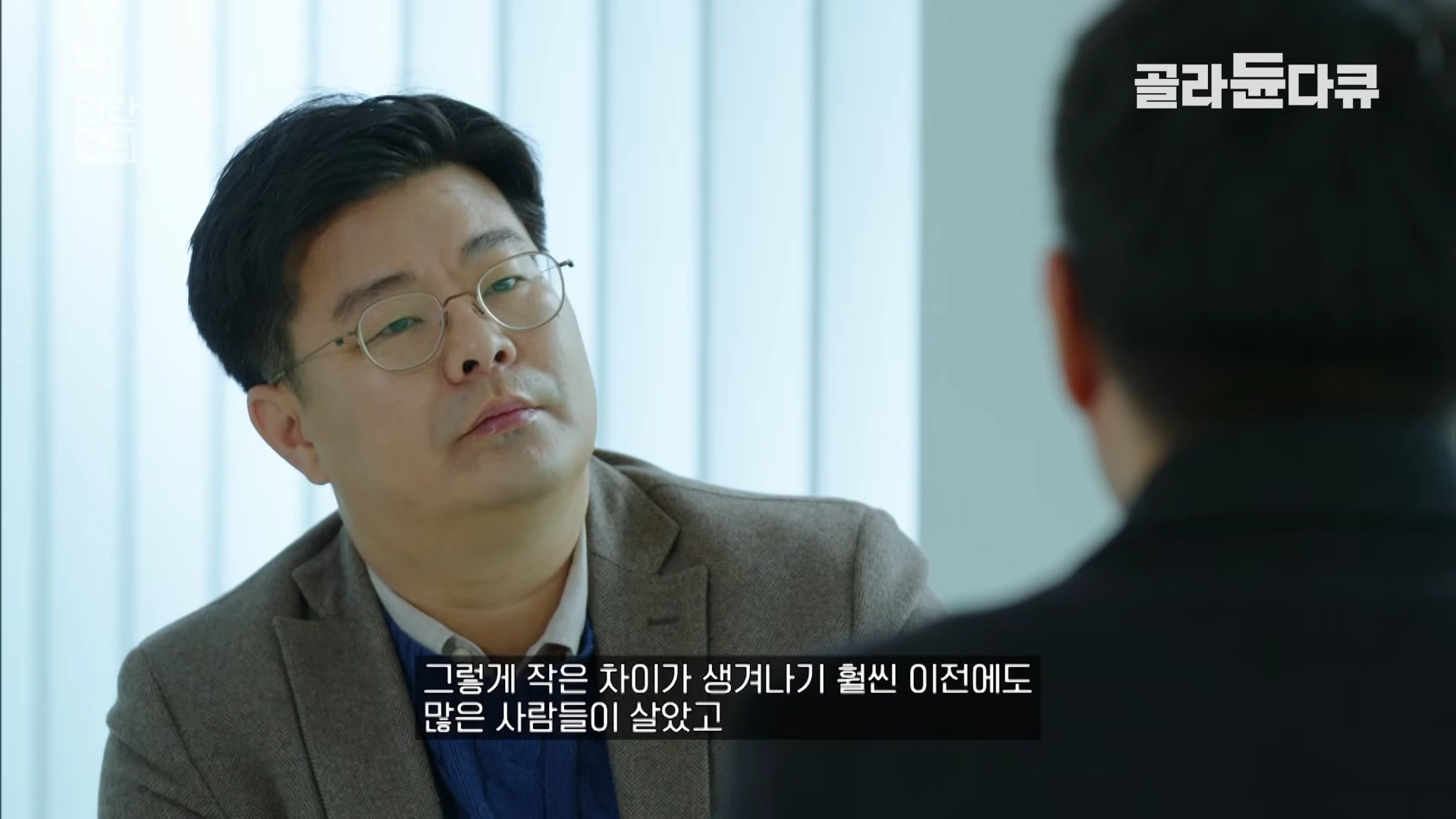 안경을 쓴 남성이 상대방을 바라보며 대화하는 모습이 담긴 다큐멘터리 영상 화면