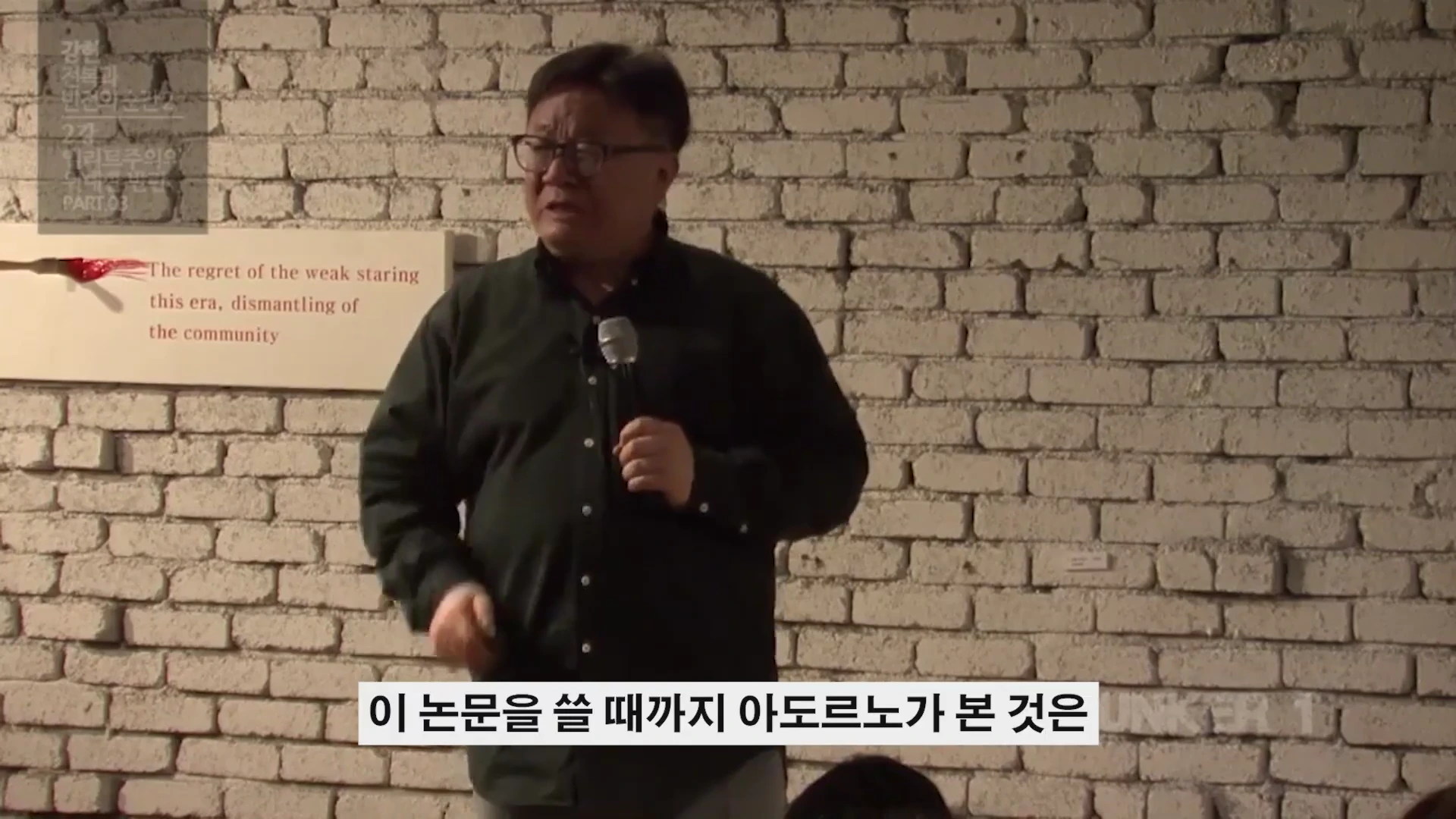 흰 벽돌 배경 앞에서 마이크를 들고 강연하는 중년 남성