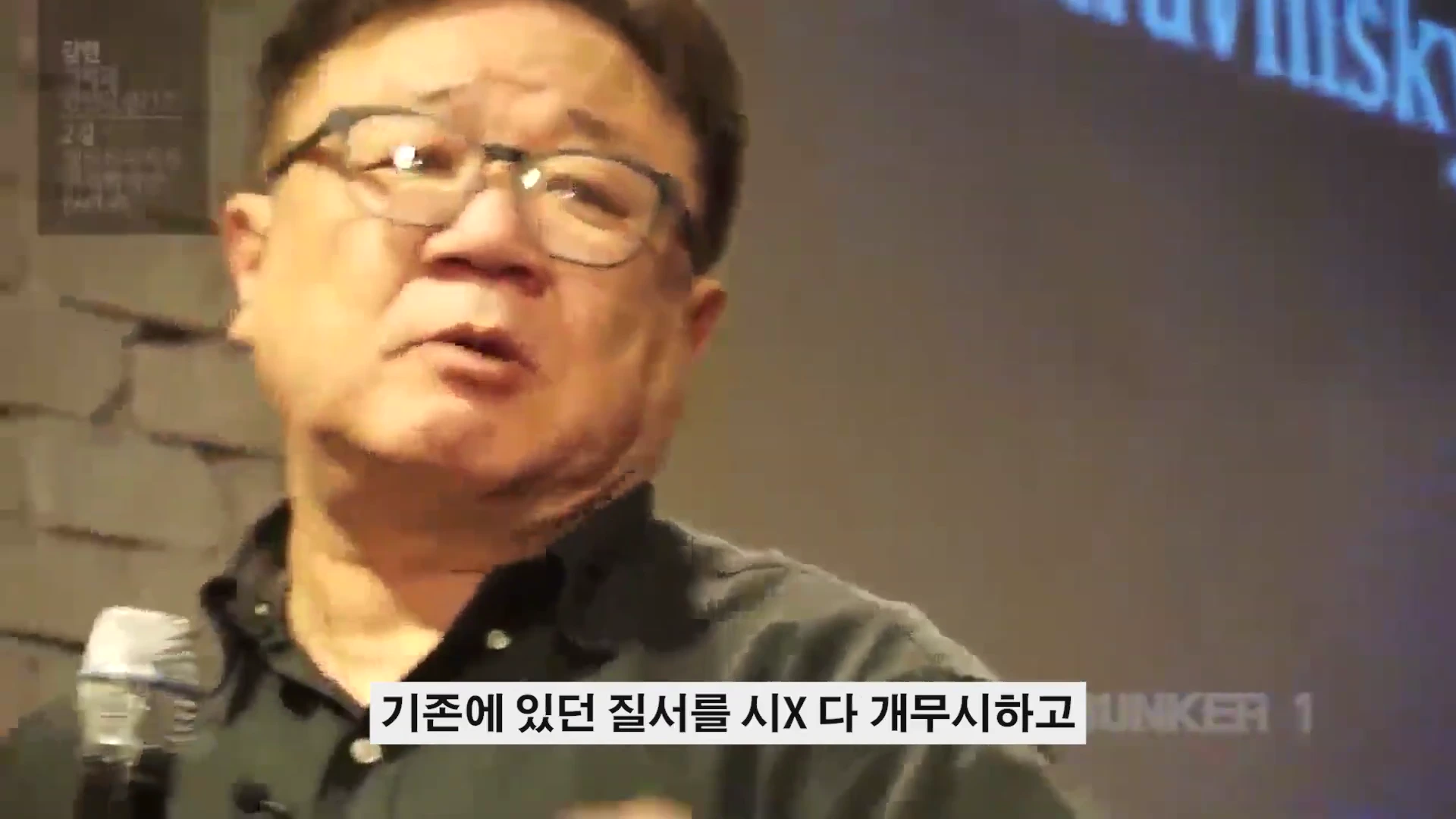 안경을 쓴 중년 남성이 강연장에서 마이크를 들고 열정적으로 이야기하는 모습