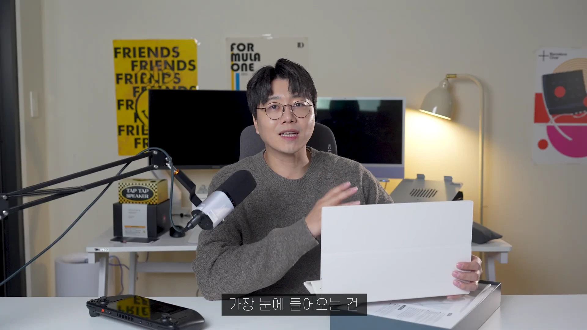 안경을 쓴 남성이 책상에 앉아 하얀색 포터블 모니터를 손에 들고 설명하는 모습