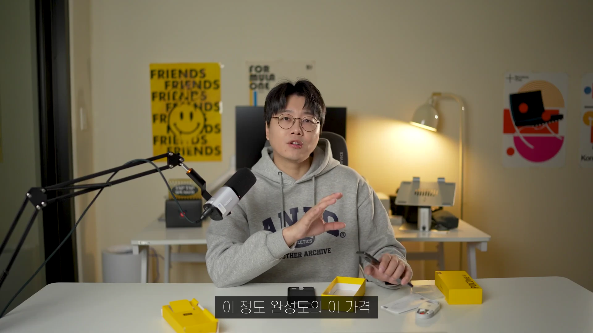 안경을 쓴 남성이 책상에 앉아 스마트폰을 손에 들고 설명하는 모습