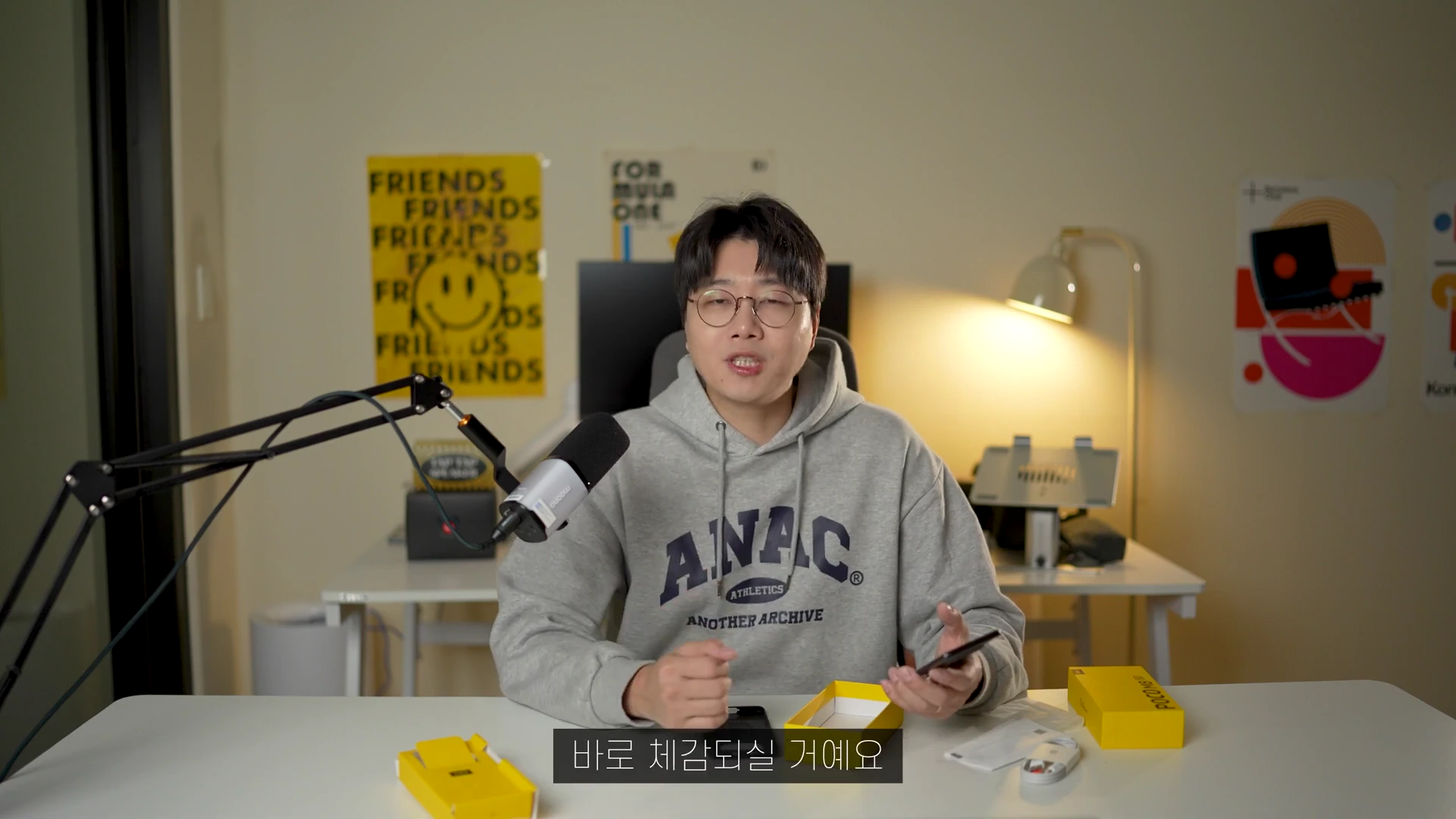안경을 쓴 남성이 책상에 앉아 스마트폰을 들고 설명하는 모습