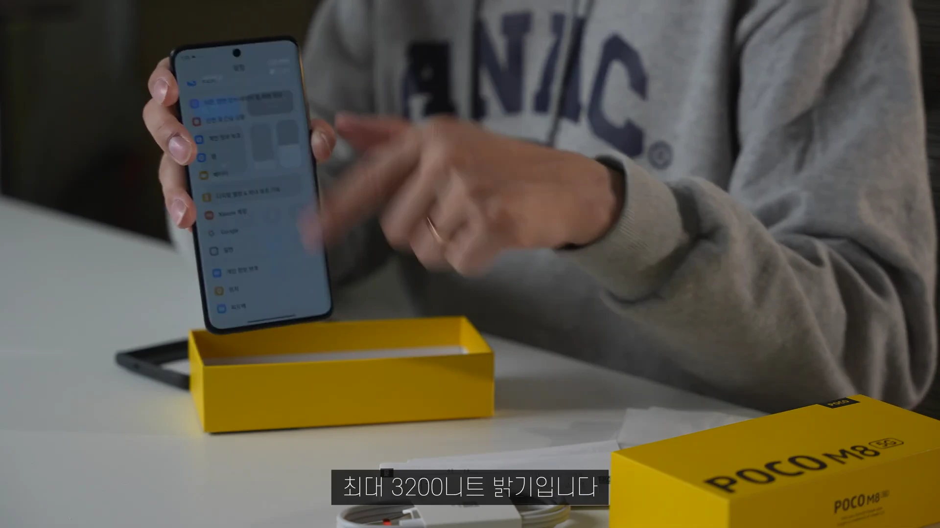 한 사람이 손에 스마트폰을 들고 화면을 가리키고 있으며, 앞에는 노란색 제품 상자가 놓여 있다.