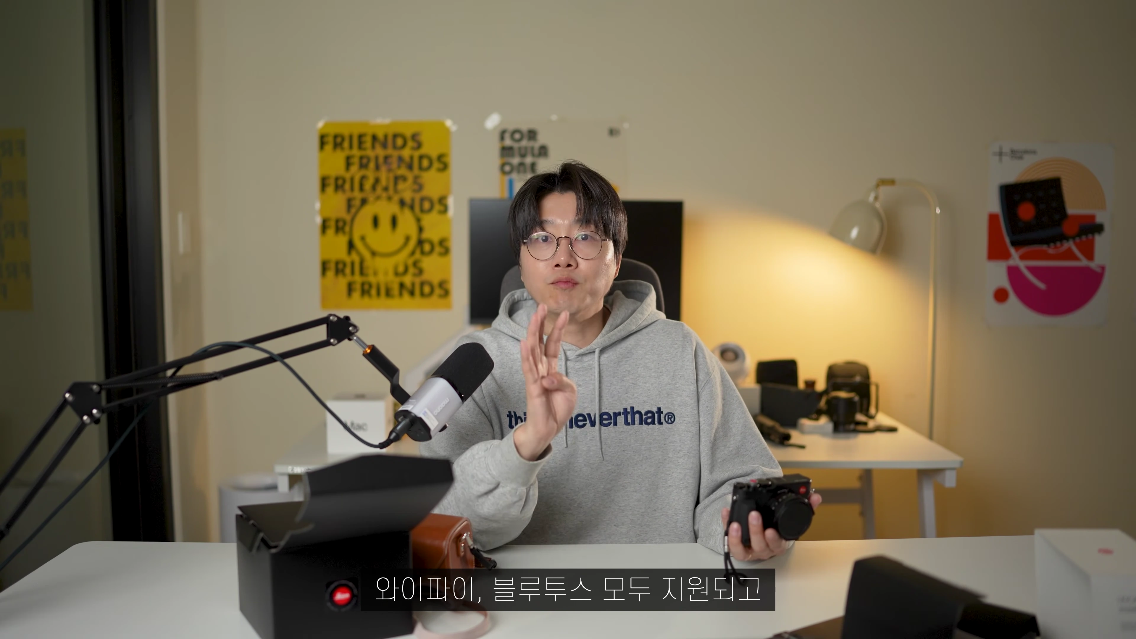 안경을 쓴 남성이 회색 후드티를 입고 책상 앞에 앉아 카메라를 들고 설명하는 모습