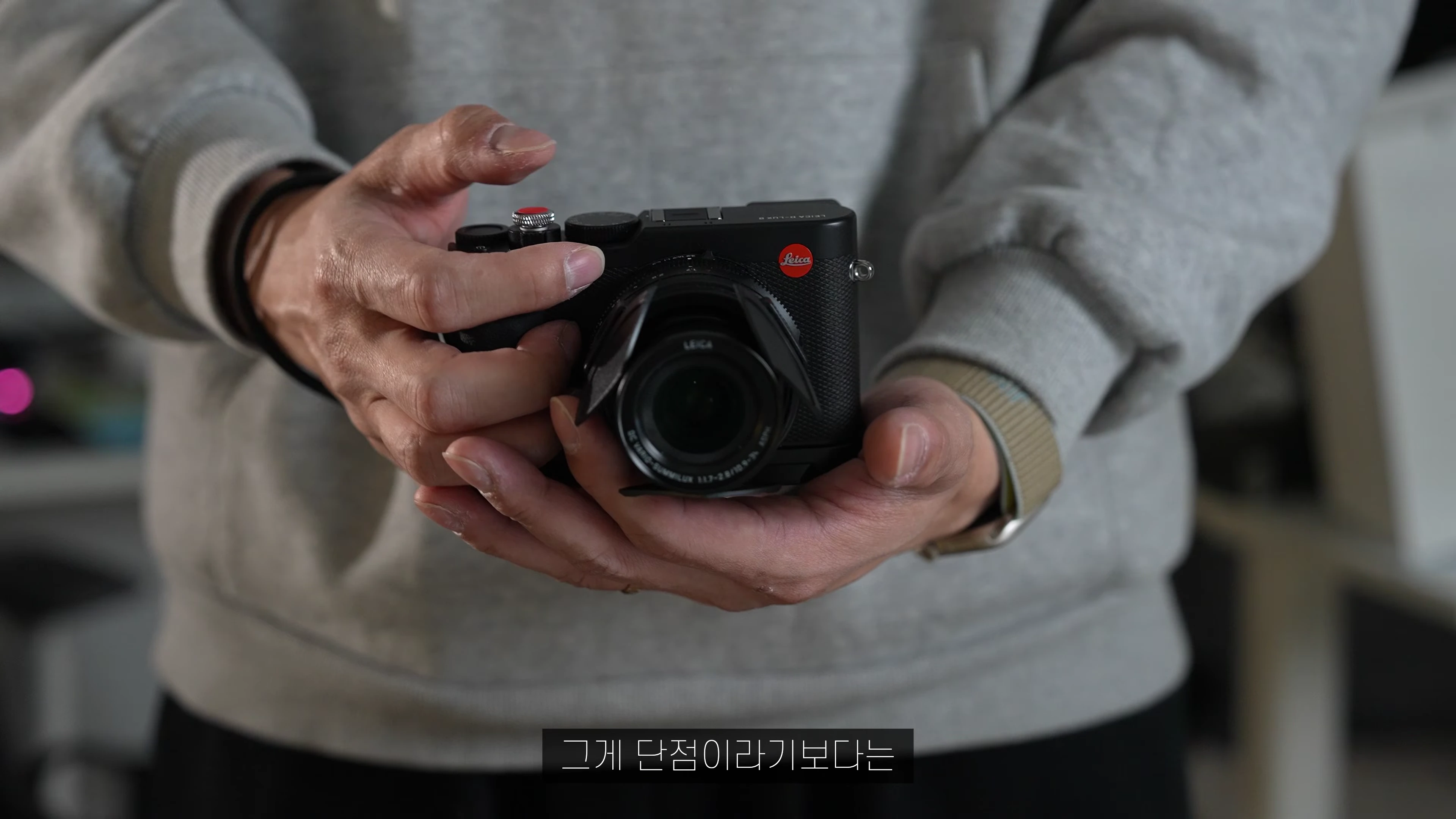 회색 후드를 입은 사람이 검은색 라이카 카메라를 양손으로 들고 조작하는 모습