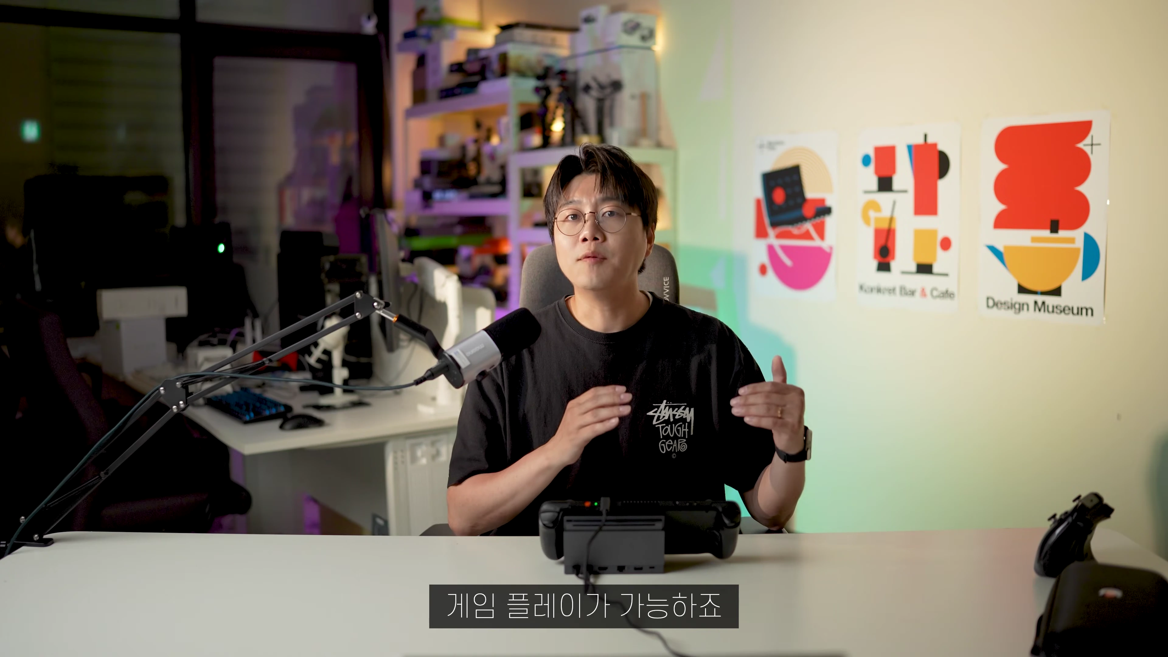 안경을 쓴 남성이 책상 앞에 앉아 스팀덱이 연결된 도킹 스테이션을 앞에 두고 설명하고 있다.