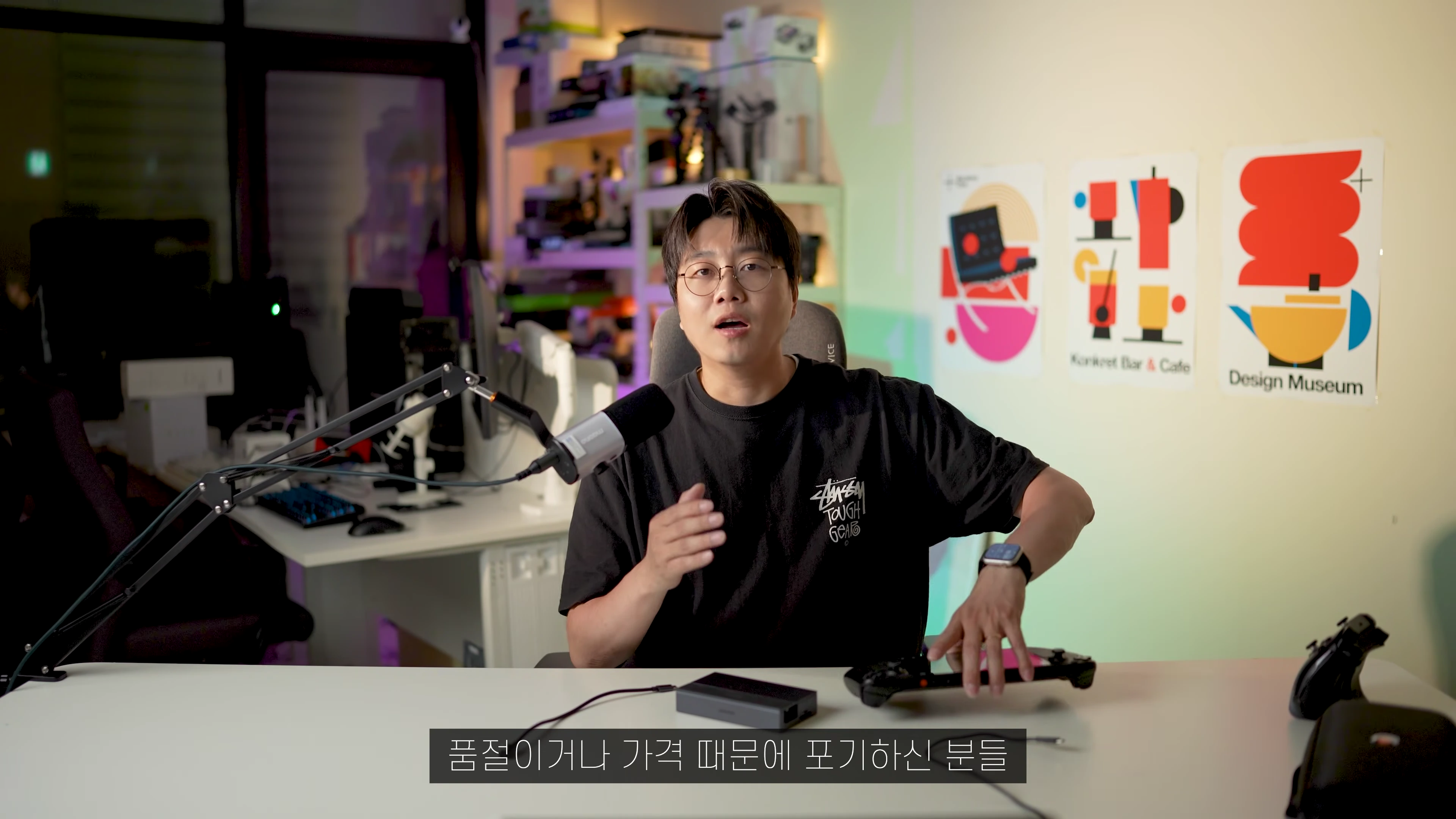 안경을 쓴 남성이 책상 앞에 앉아 스팀덱과 도킹 스테이션을 소개하고 있는 모습