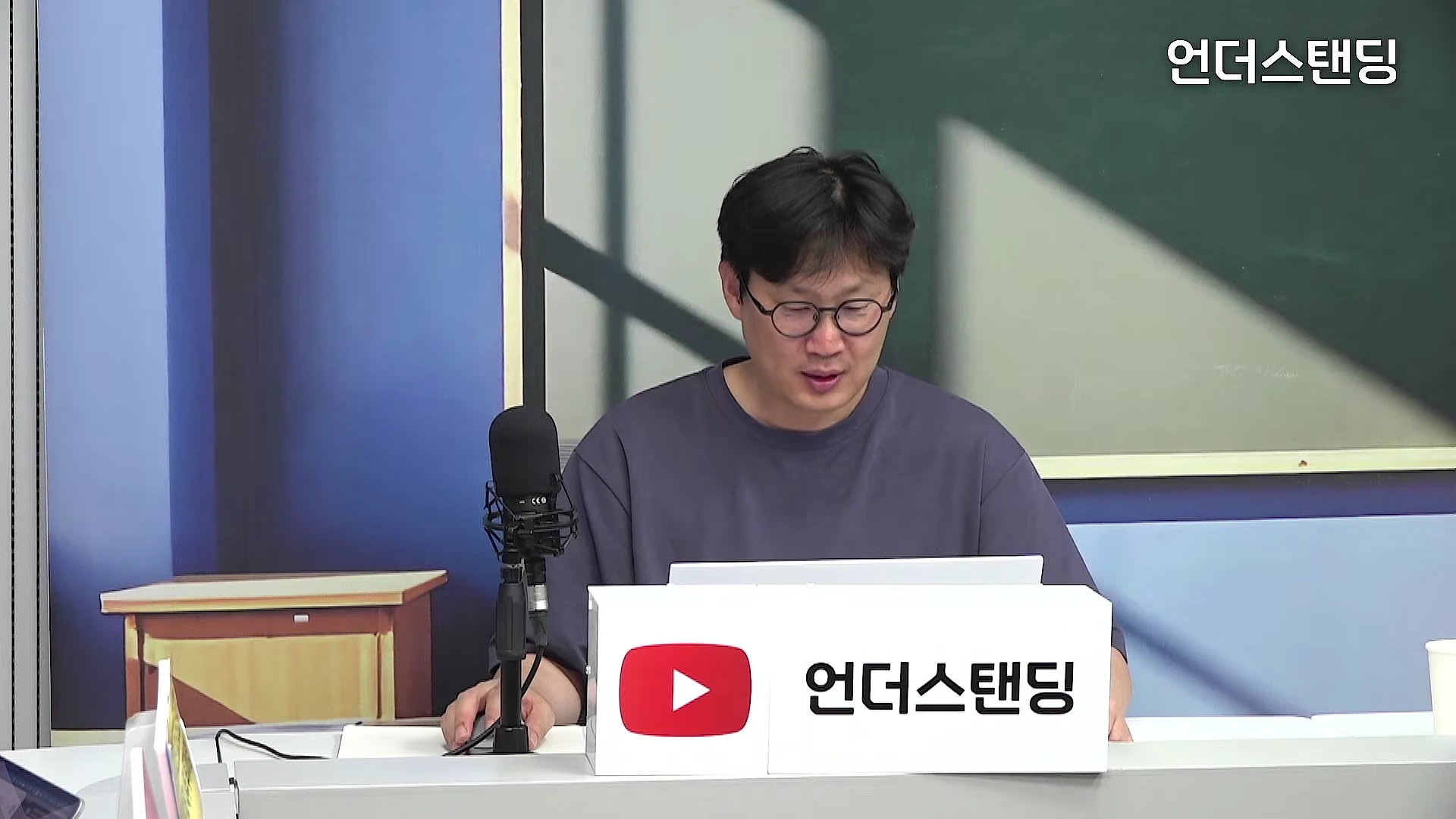 안경을 쓴 남성이 스튜디오 책상 앞에 앉아 노트북을 보며 이야기하는 모습