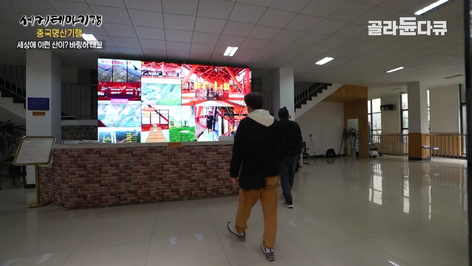 실내 로비 벽면에 설치된 대형 모니터에 바링허 대교의 모습이 여러 화면으로 분할되어 나오고 있고, 그 앞을 두 사람이 걸어가고 있다.