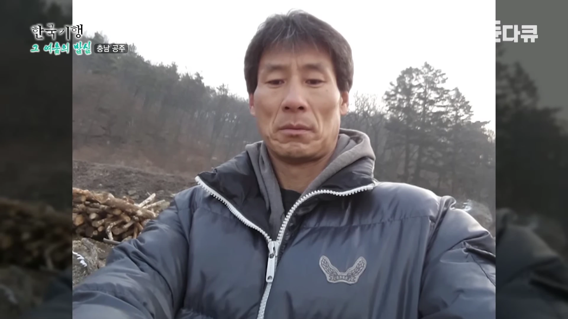 겨울 산을 배경으로 두꺼운 패딩 점퍼를 입고 서 있는 중년 남성의 모습