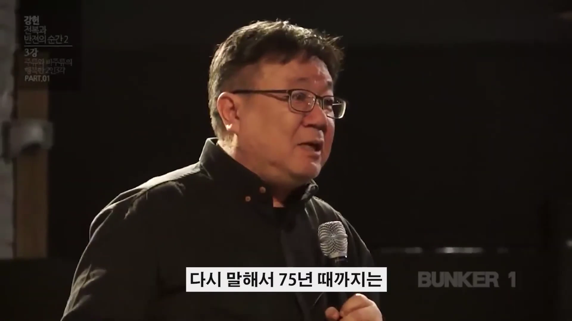 안경을 쓴 중년 남성이 어두운 무대 위에서 마이크를 잡고 강연하는 모습