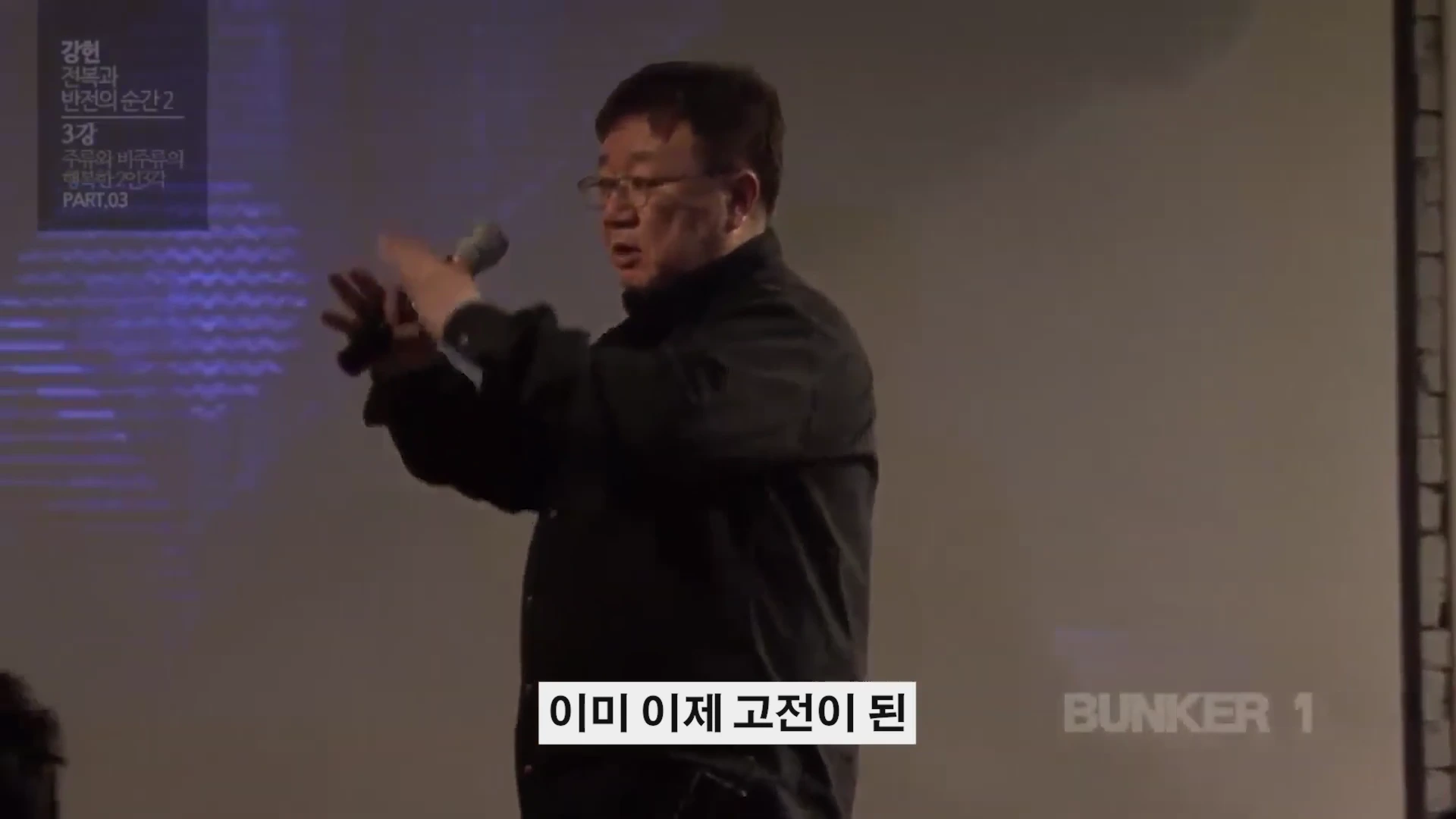 안경을 쓴 중년 남성이 마이크를 들고 강연장에서 손짓하며 열정적으로 이야기하고 있다.