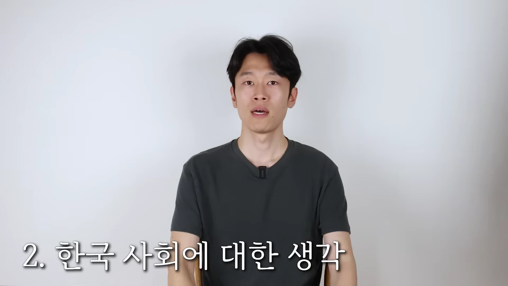 흰 배경 앞에 앉아 카메라를 정면으로 응시하며 이야기하는 남성