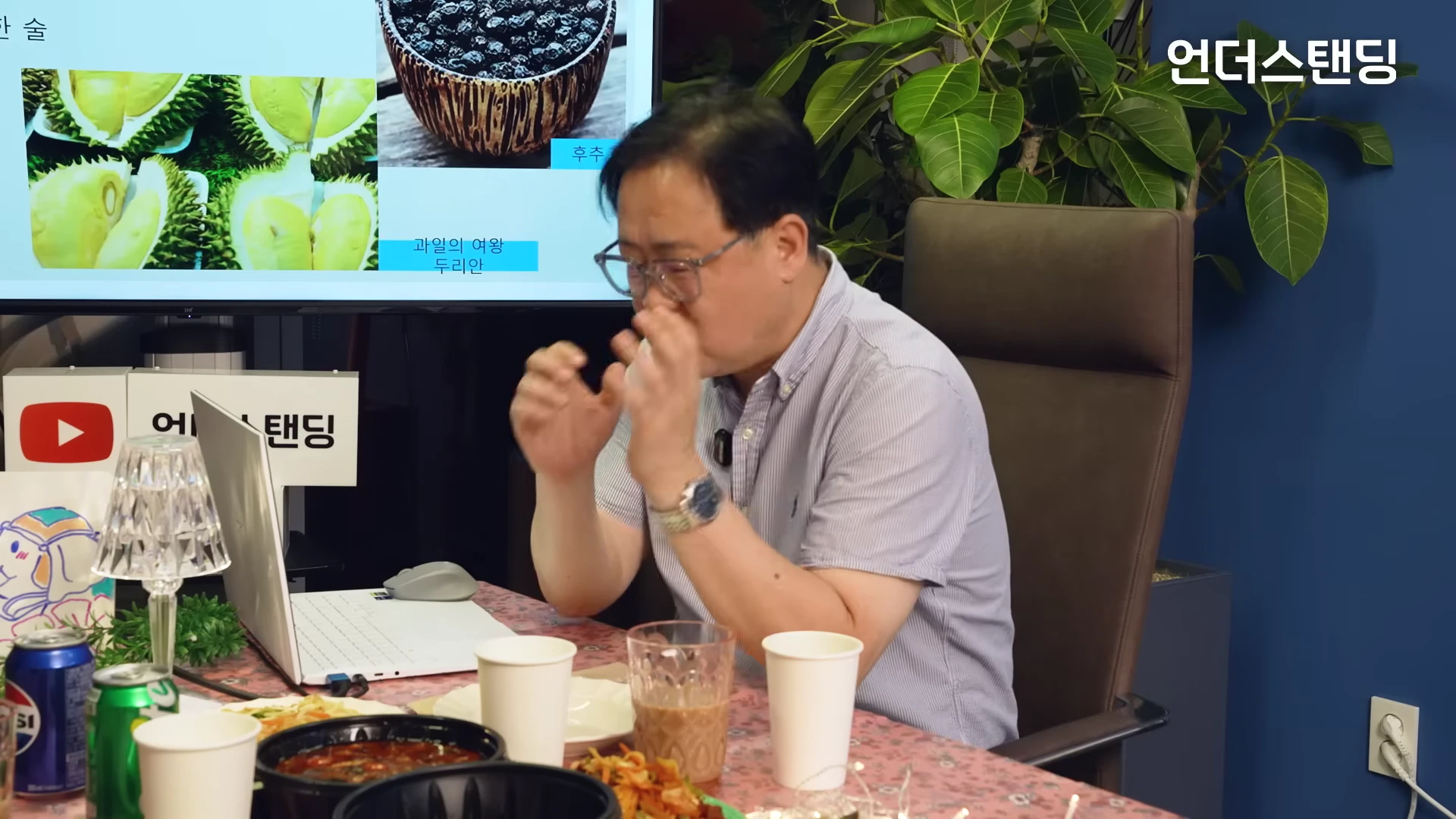스튜디오에서 안경을 쓴 남성이 손짓하며 이야기하고 있고, 뒤편 화면에는 두리안 사진이 띄워져 있다.