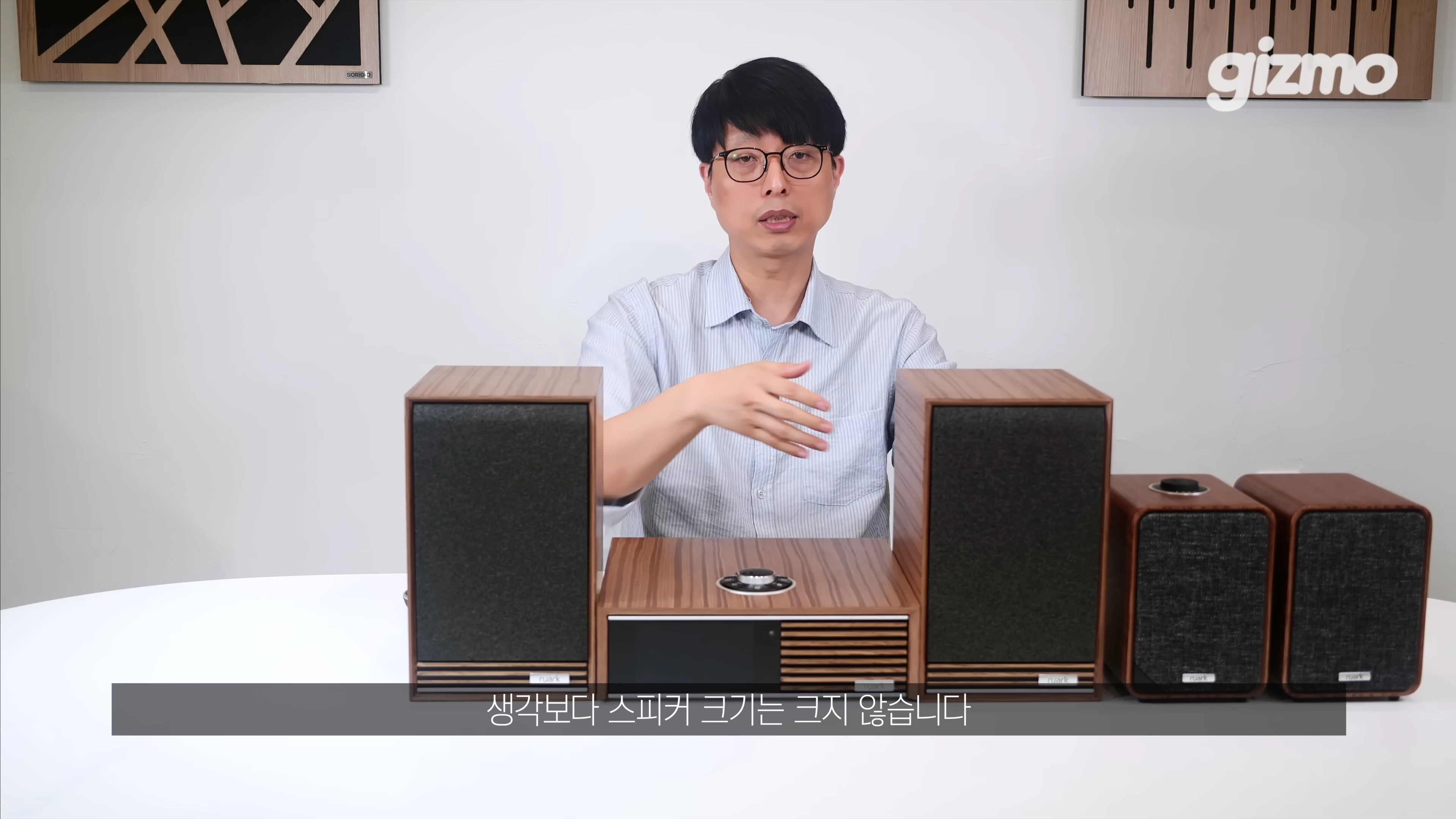 안경을 쓴 남성이 테이블 위에 놓인 루악 오디오 시스템과 스피커를 소개하고 있다.