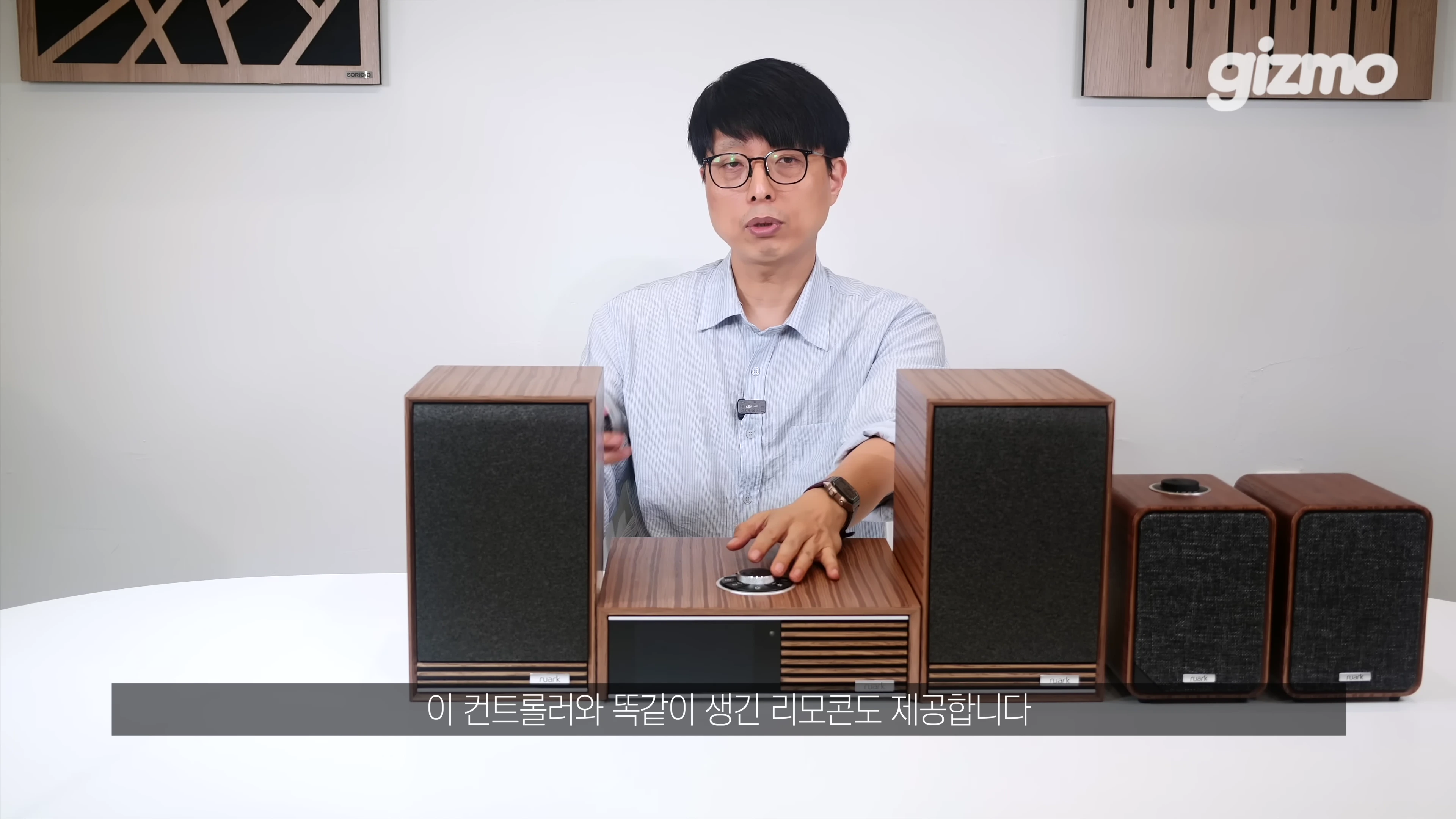 안경을 쓴 남성이 테이블 위에 놓인 루악 오디오 본체와 스피커 세트를 손으로 가리키며 설명하고 있다.