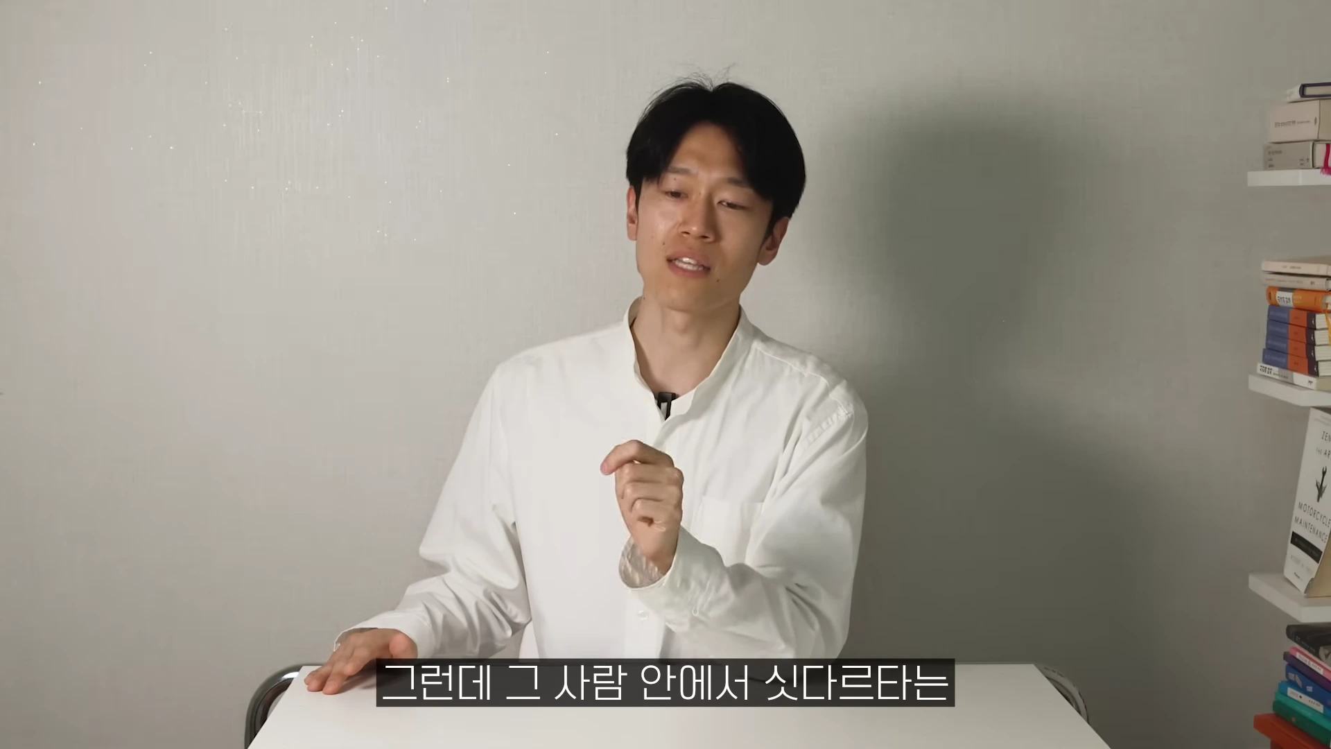 흰 셔츠를 입은 남성이 책장이 있는 방에서 손짓하며 이야기하고 있다.
